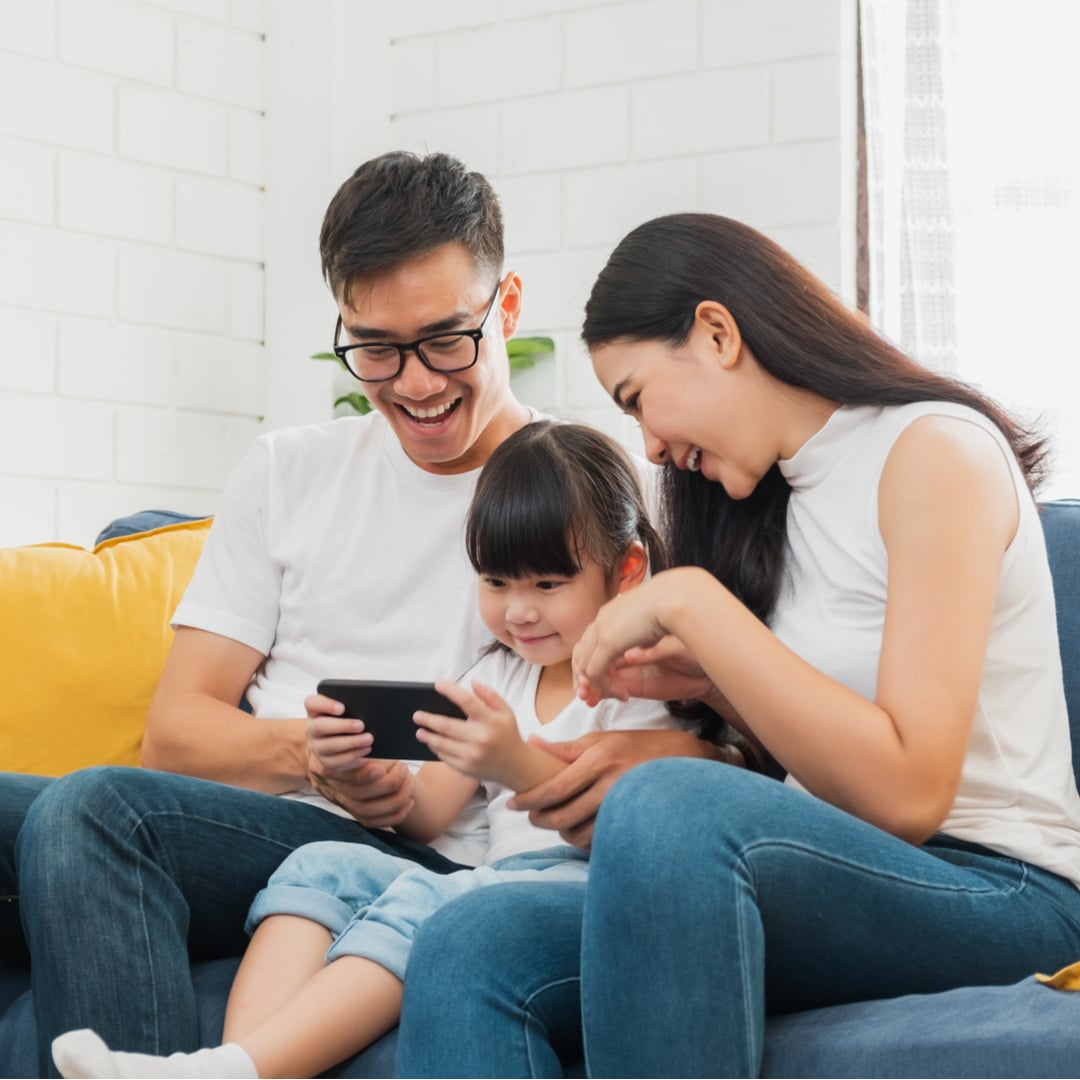 Asian American couple, with mom and dad coparenting a young child. They are watching a video together on the child's phone and have happy expressions on their faces.