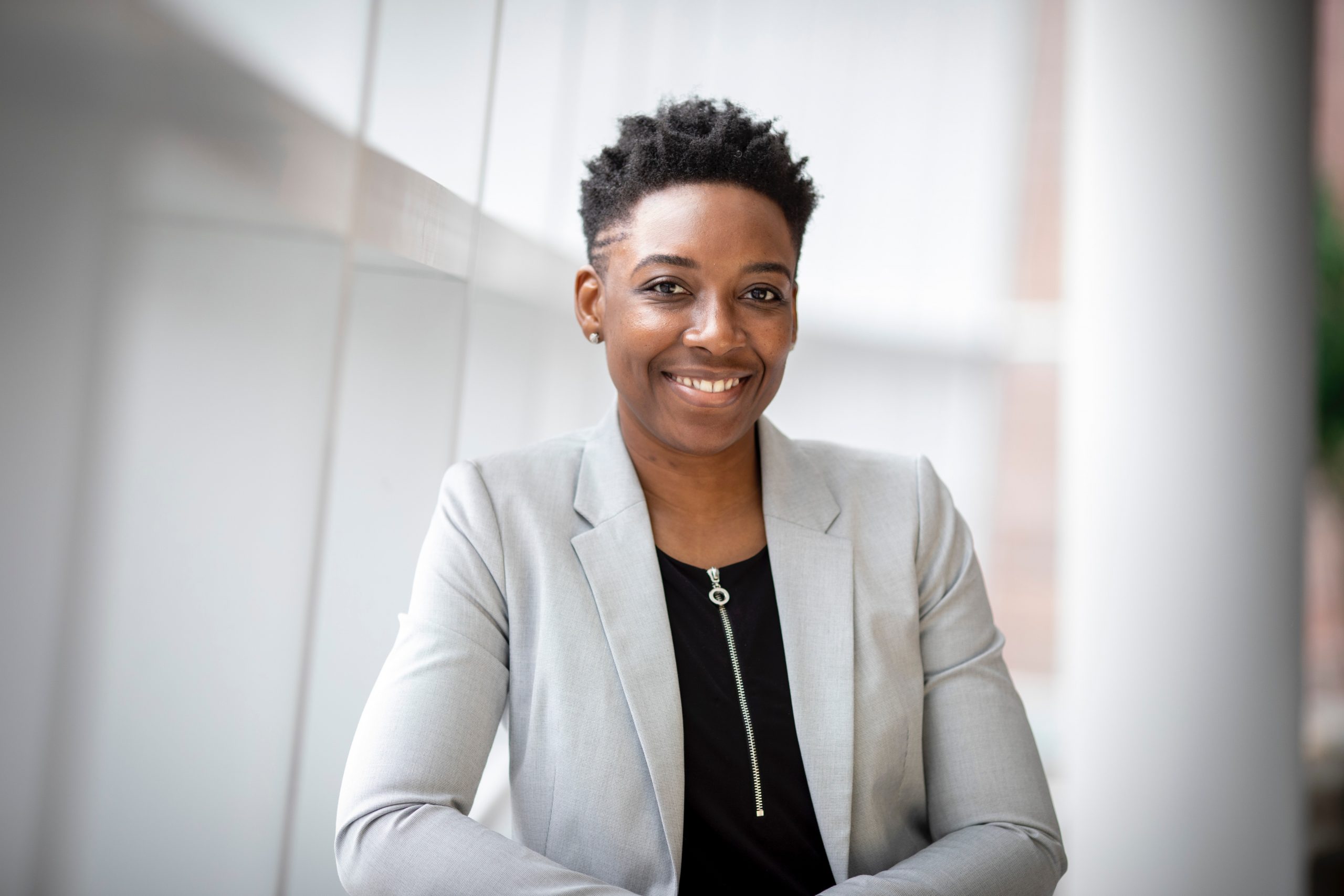 Young Black woman with short natural hair smiles at the camera. She is wearing a light gray blazer and black blouse with a zipper.