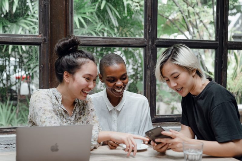 A group of three young professionals working together with happy, relaxed expressions on their face. The person on the left is a femme presenting Asian American with long hair tied back. The middle person is an androgynous Black person with shaved head and a button down shirt. The person on the right is an androgynous Asian person with short, bleached hair wearing a black t-shirt.