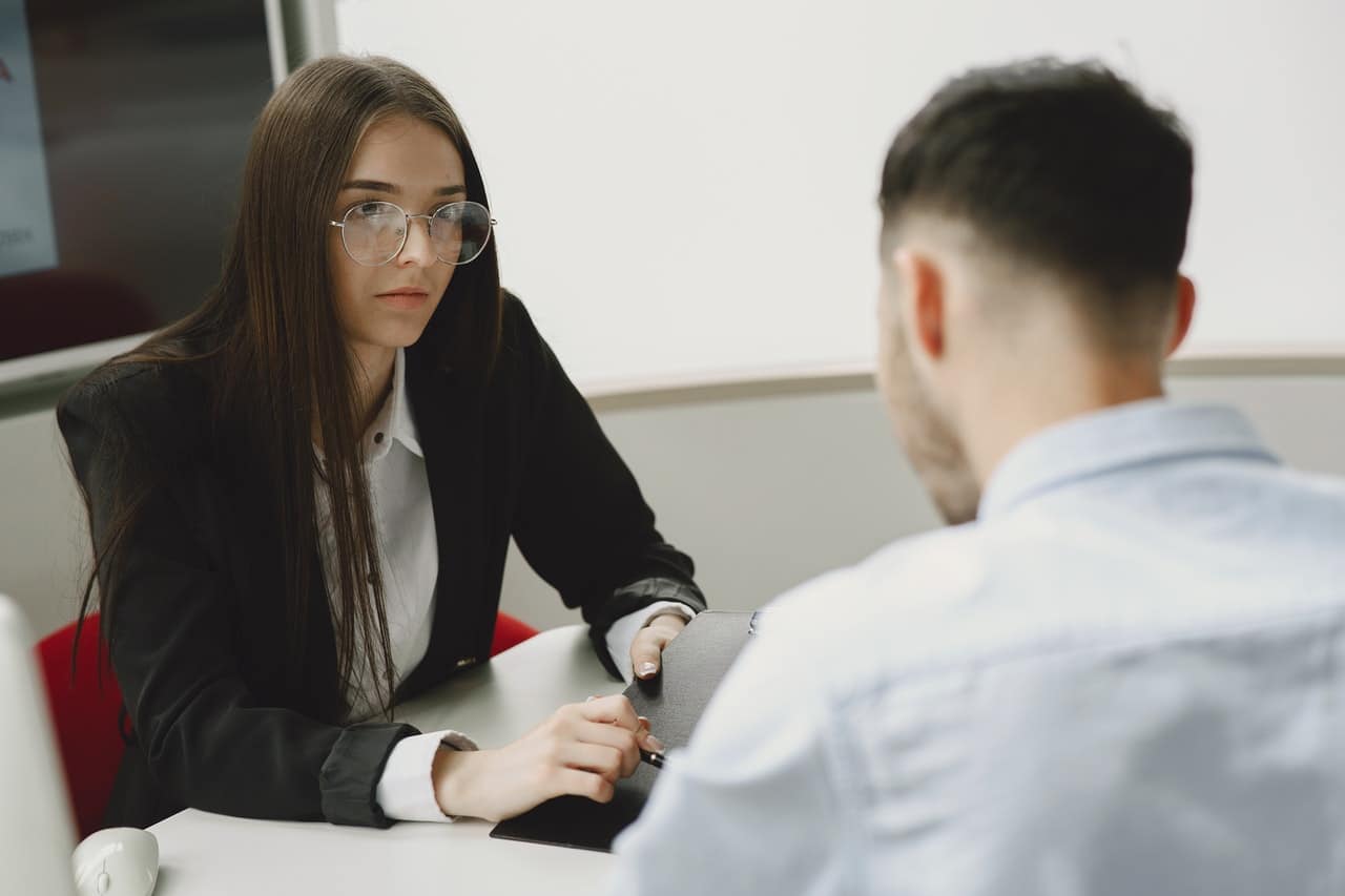 A long haired woman is sitting at a desk across from a man. They are both in professional attired and she has a serious look on her face.