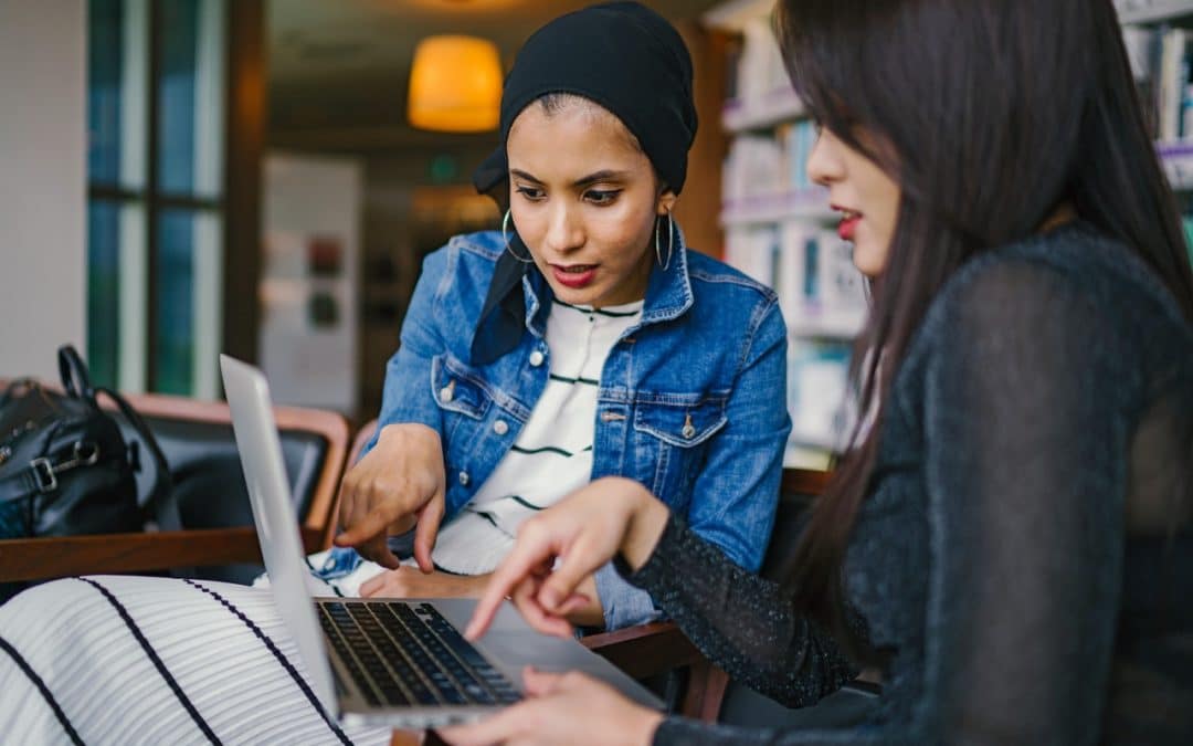 Two Millennial women looking at a laptop together. The first is a brown skinned women with a head scarf and hoop earrings. The second is an Asian American woman with long hair and red lipstick.