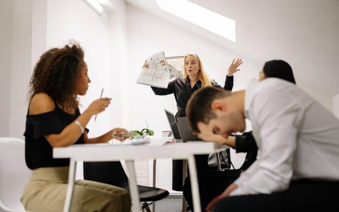 Workers in an office looking stressed and upset. A long haired blonde woman appears to be throwing her hands up in frustration at the team. A man in the foreground looks down at his feet in a hunched over position.