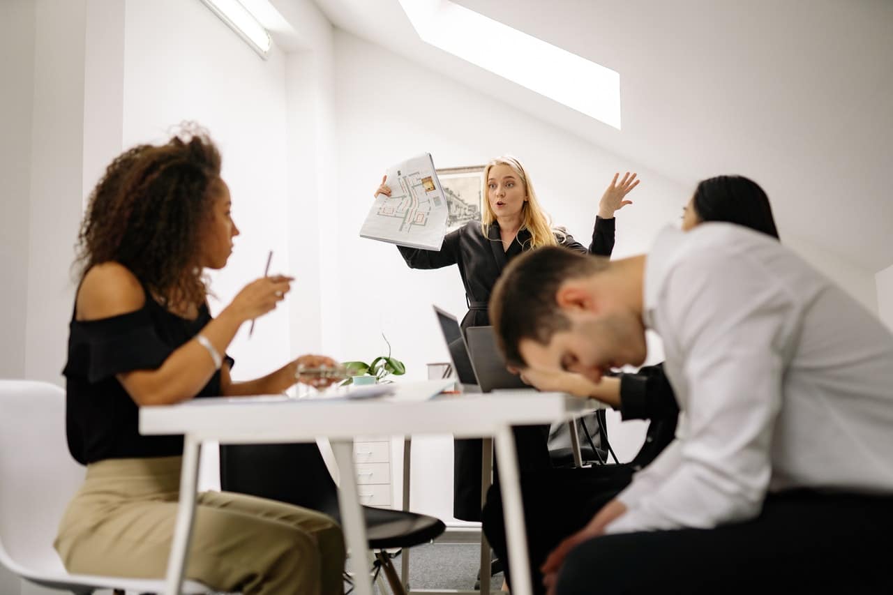 Workers in an office looking stressed and upset. A long haired blonde woman appears to be throwing her hands up in frustration at the team. A man in the foreground looks down at his feet in a hunched over position.