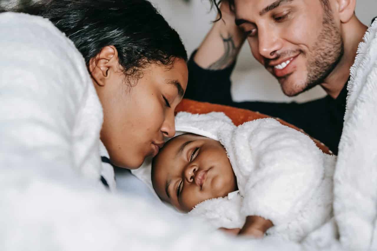 Two parents cuddling a young baby together. One is kissing the baby on their forehead.