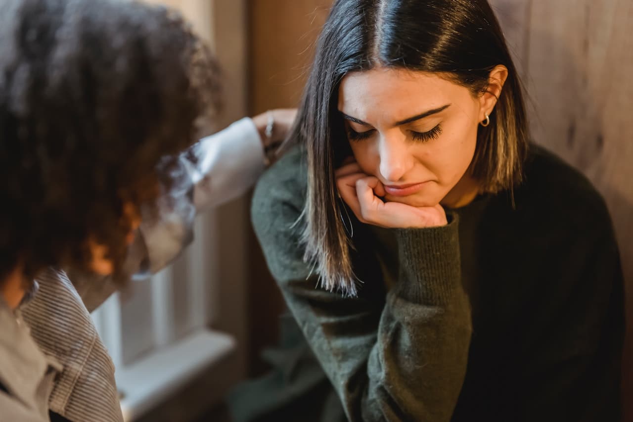 A sad looking woman with a dark sweater and shoulder length hair looks down. A second person is partially shown in the foreground and is reaching out to her.