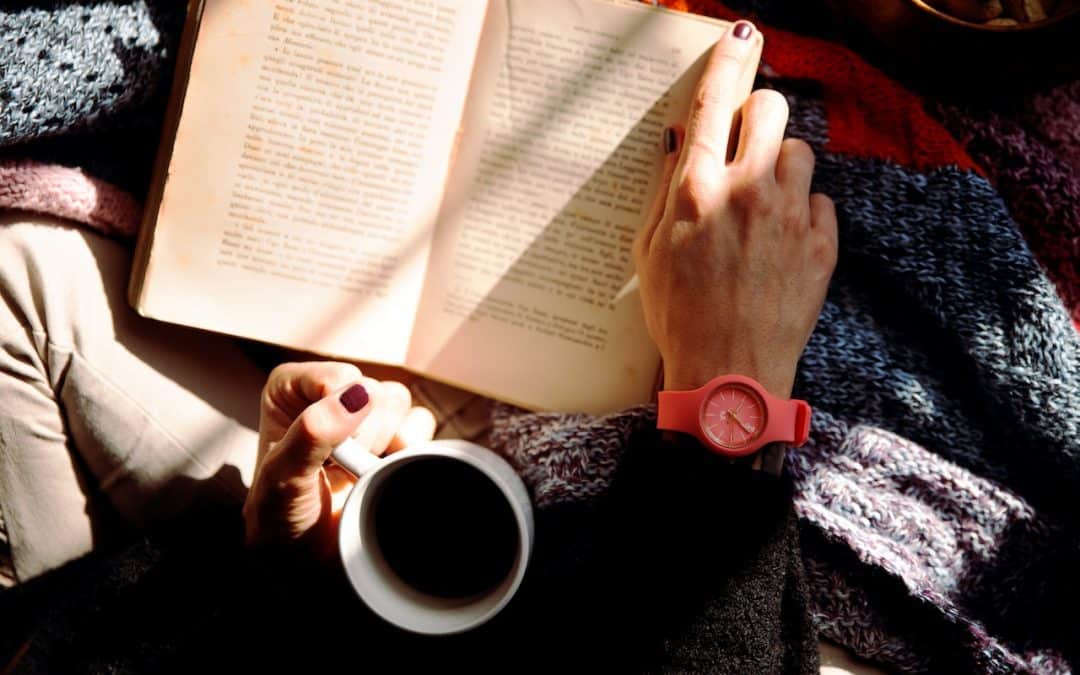 Photo of a person reading a book and drinking coffee from above. Only their hands and forearms are visible.