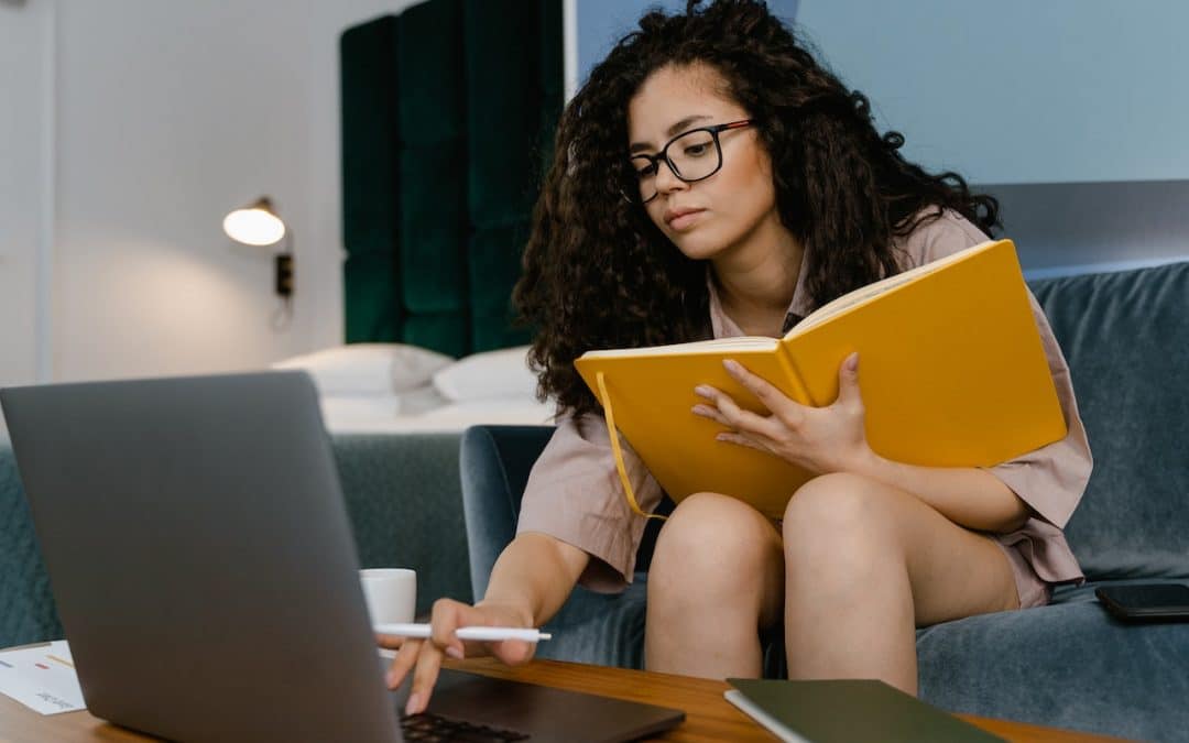 A curly haired woman leans over a book while working on her laptop