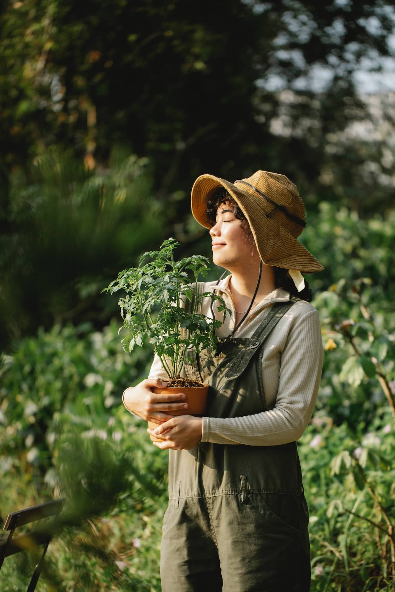 An Asian American woman carrying a potted plant outdoors with a happy look on her face. She is dressed in overalls and wearing a sun hat.