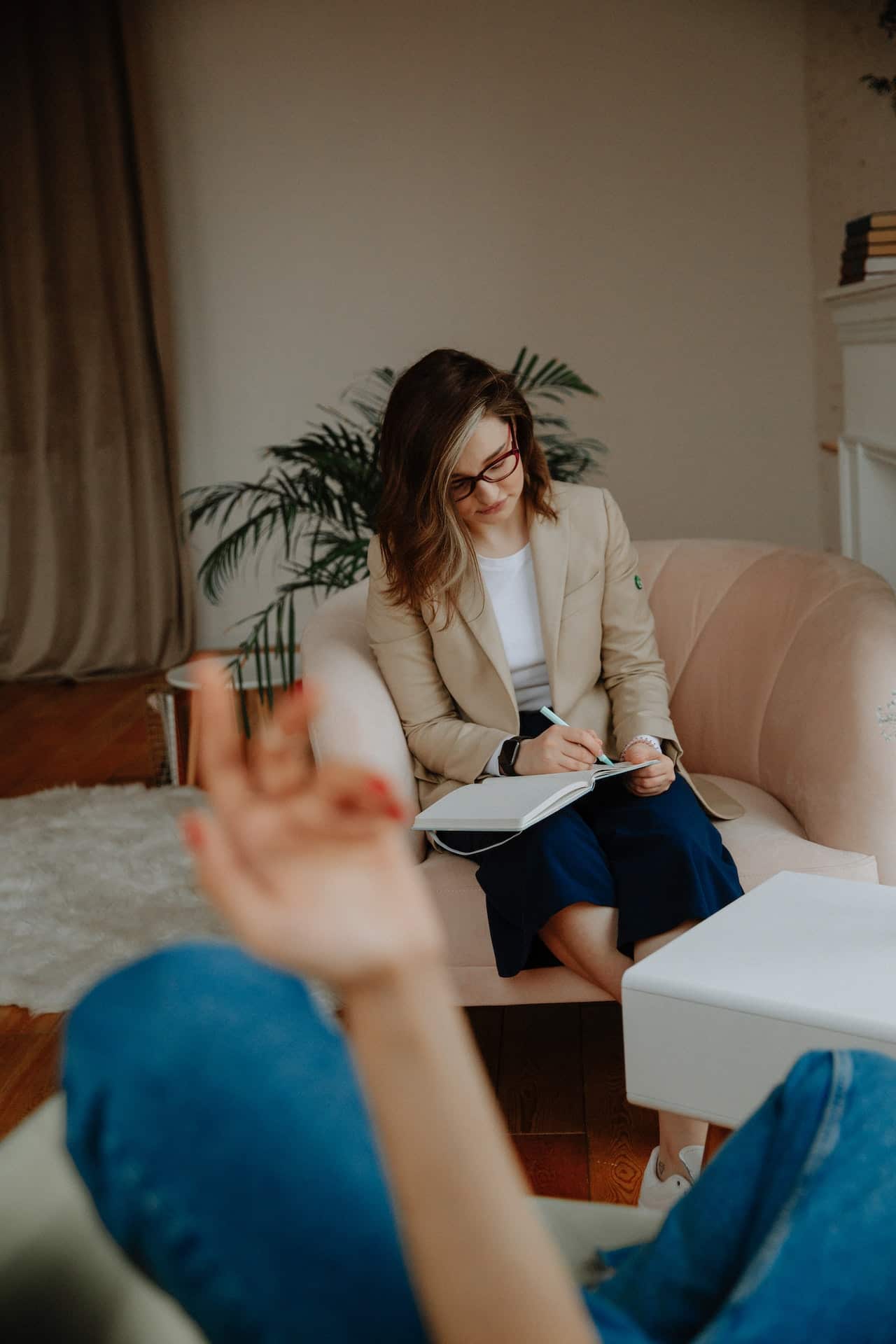 Therapist in a blazer and dark pants writes in a notebook. Client can be seen in corner of the foreground.