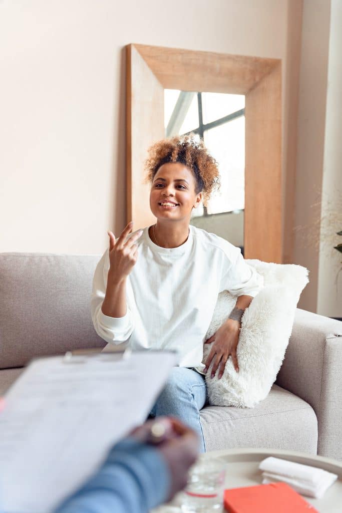 A Black woman with curly hair, dressed casually, sits on a therapy couch with a smiling look on her face. A therapist's arm and notebook is visible in the foreground.