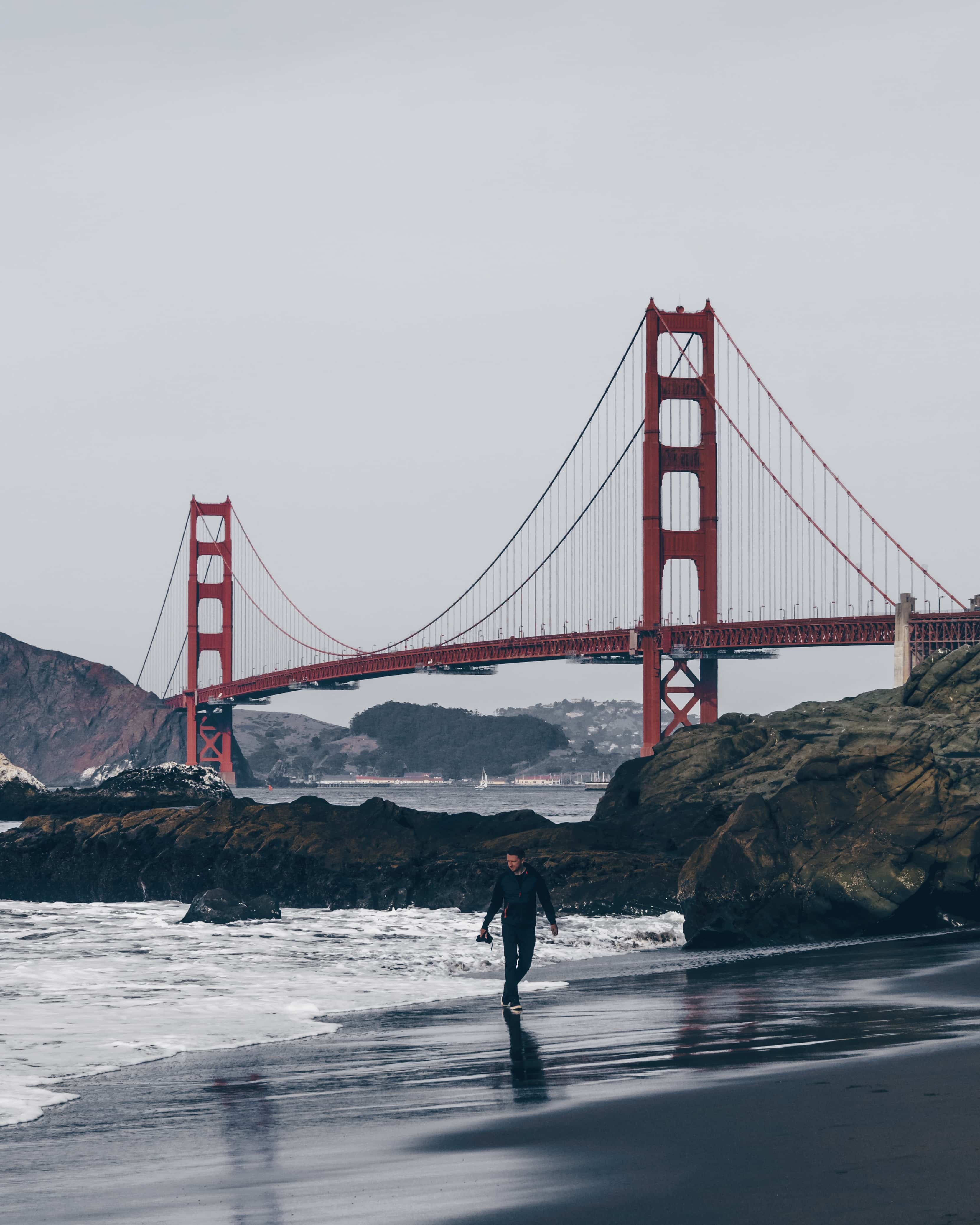 Golden Gate Bridge in San Francisco photographed from Baker Beach on a cloudy day. A person is standing in the distance in front of the bridge.