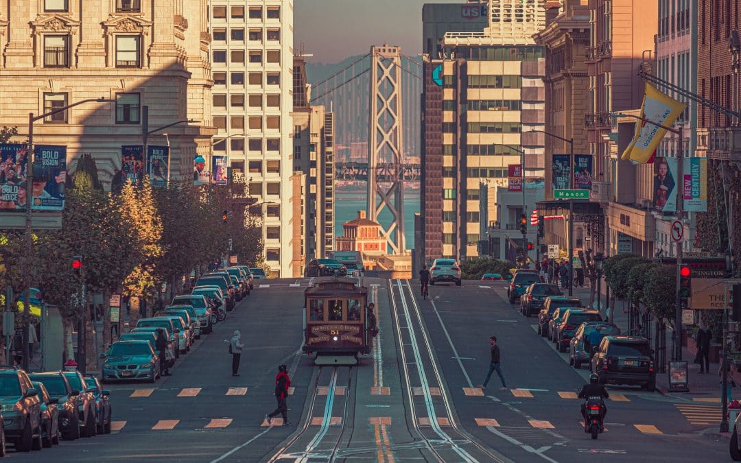 View of a San Francisco street with a cable car, a view of the Bay Bridge seen between the buildings in the distance.