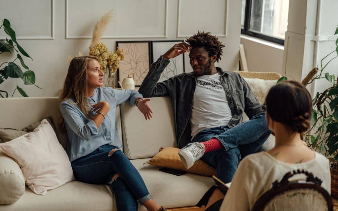 Couple in therapy sitting on a couch and discussing something in a relaxed manner. A couples counselor can be seen from the back in the foreground.