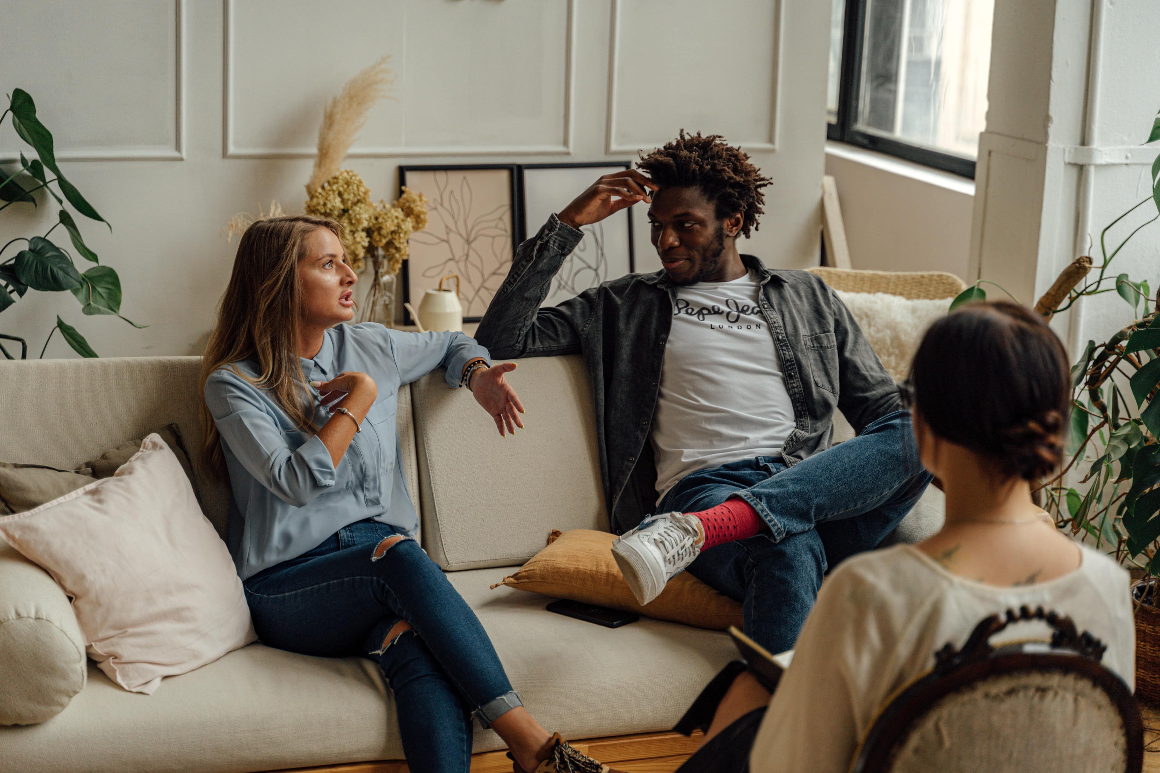 Couple in therapy sitting on a couch and discussing something in a relaxed manner. A couples counselor can be seen from the back in the foreground.