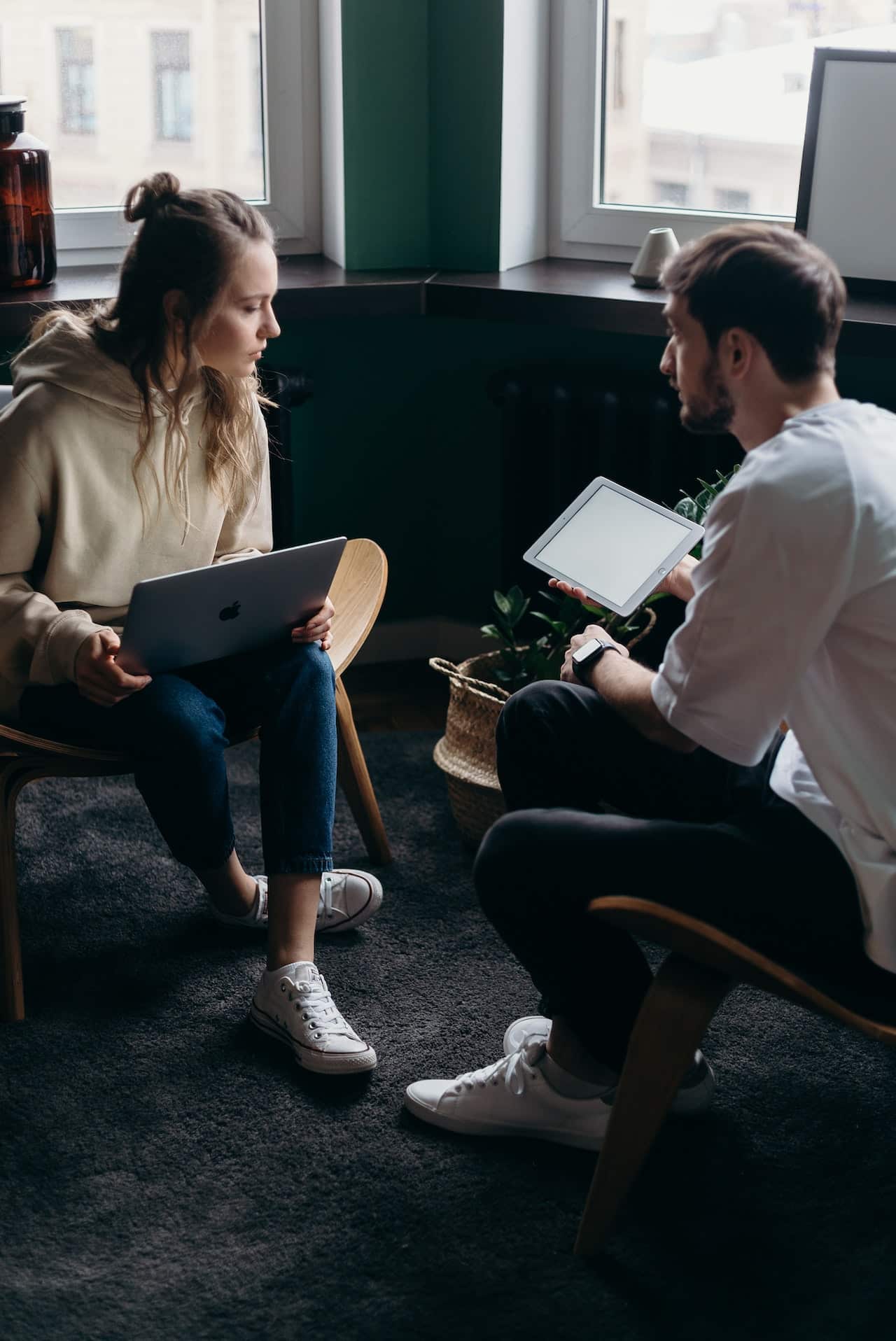 A woman sits with a laptop on her lap. A man is sitting across from her, explaining something he is showing to her on his tablet.