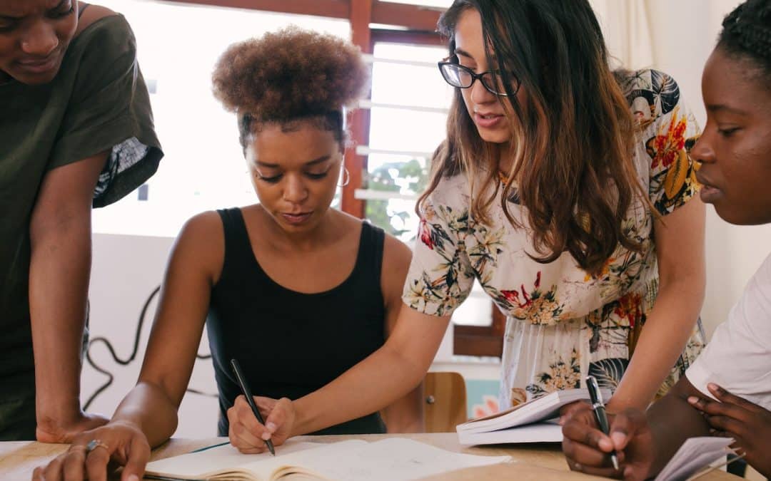 A group of women working together at a table. One is standing over the other, writing something in front of her.