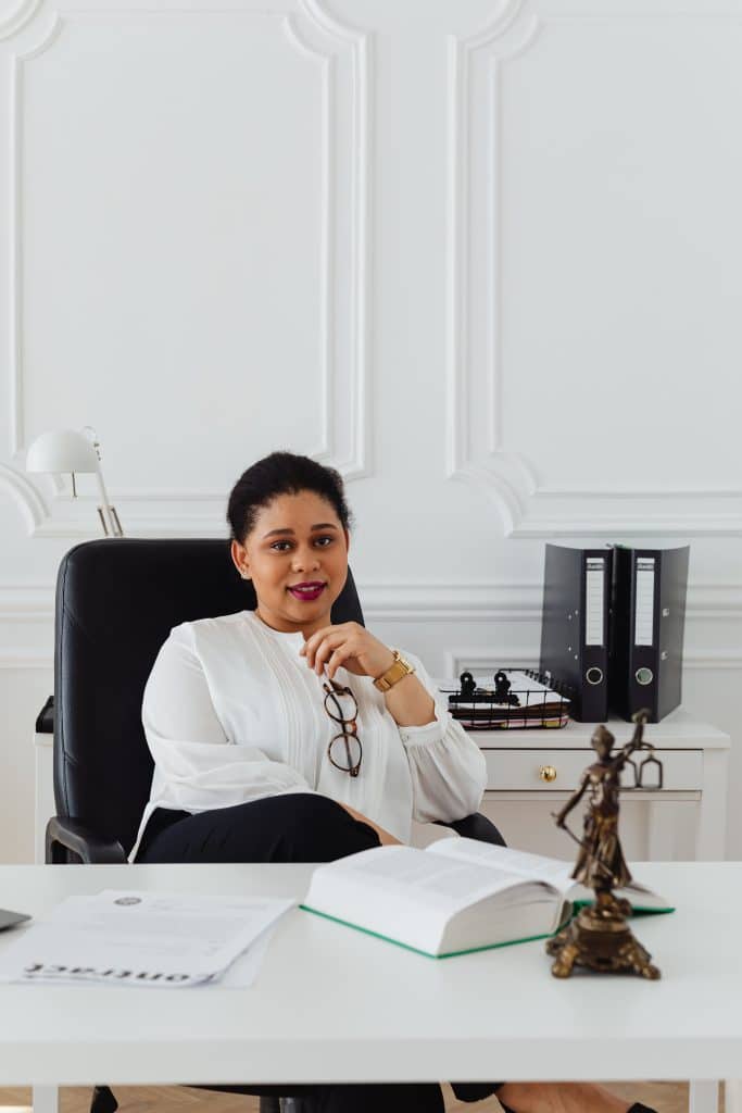 A Black professional woman sits at a desk in a white blouse and black pants, holding a pair of glasses. She looks relaxed and confident.