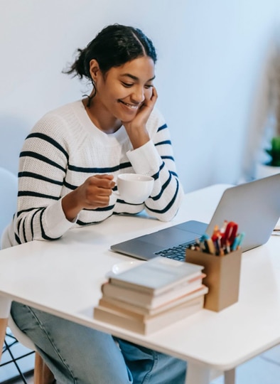 A South Asian woman working at her desk on a laptop. She is smiling and drinking a cup of coffee.