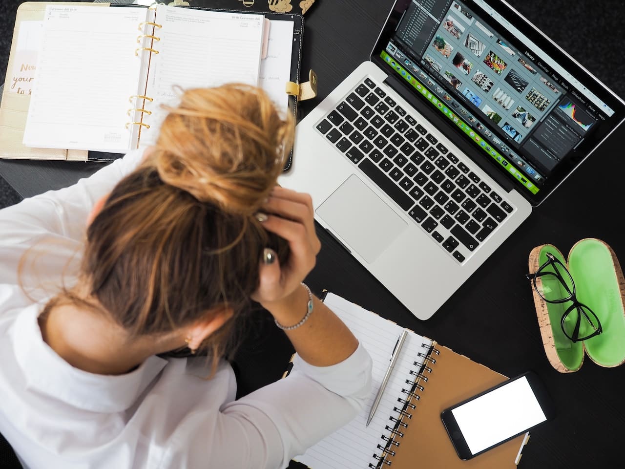 Image from above of woman with messy bun working at computer, the top of her head visible. Her hair is in a messy bun and she's holding her head in her hands.