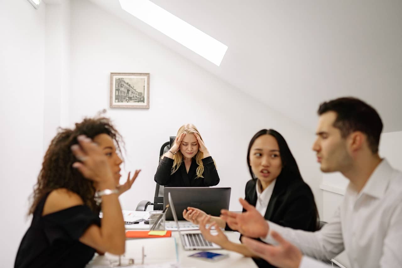 A group of people working at an office table looking stressed out and unhappy with each other.