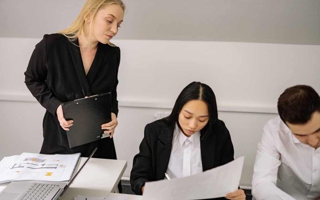 Two employees sit at work at a desk while a third looks over their shoulder with a disapproving look on their face.