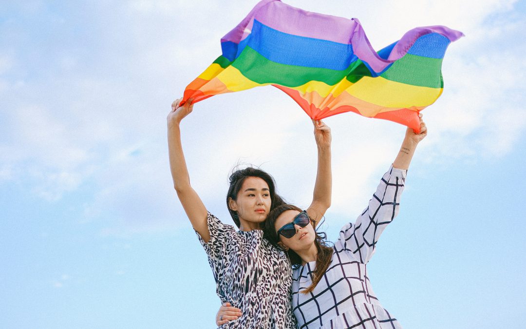 Two queer Asian American women holding a pride flag overhead, with a blue sky in the background. One has her arm around the other's waist.