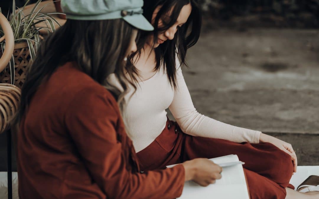 Two women, one Black and one not Black, sitting outside together, looking at a notebook on their laps, talking.