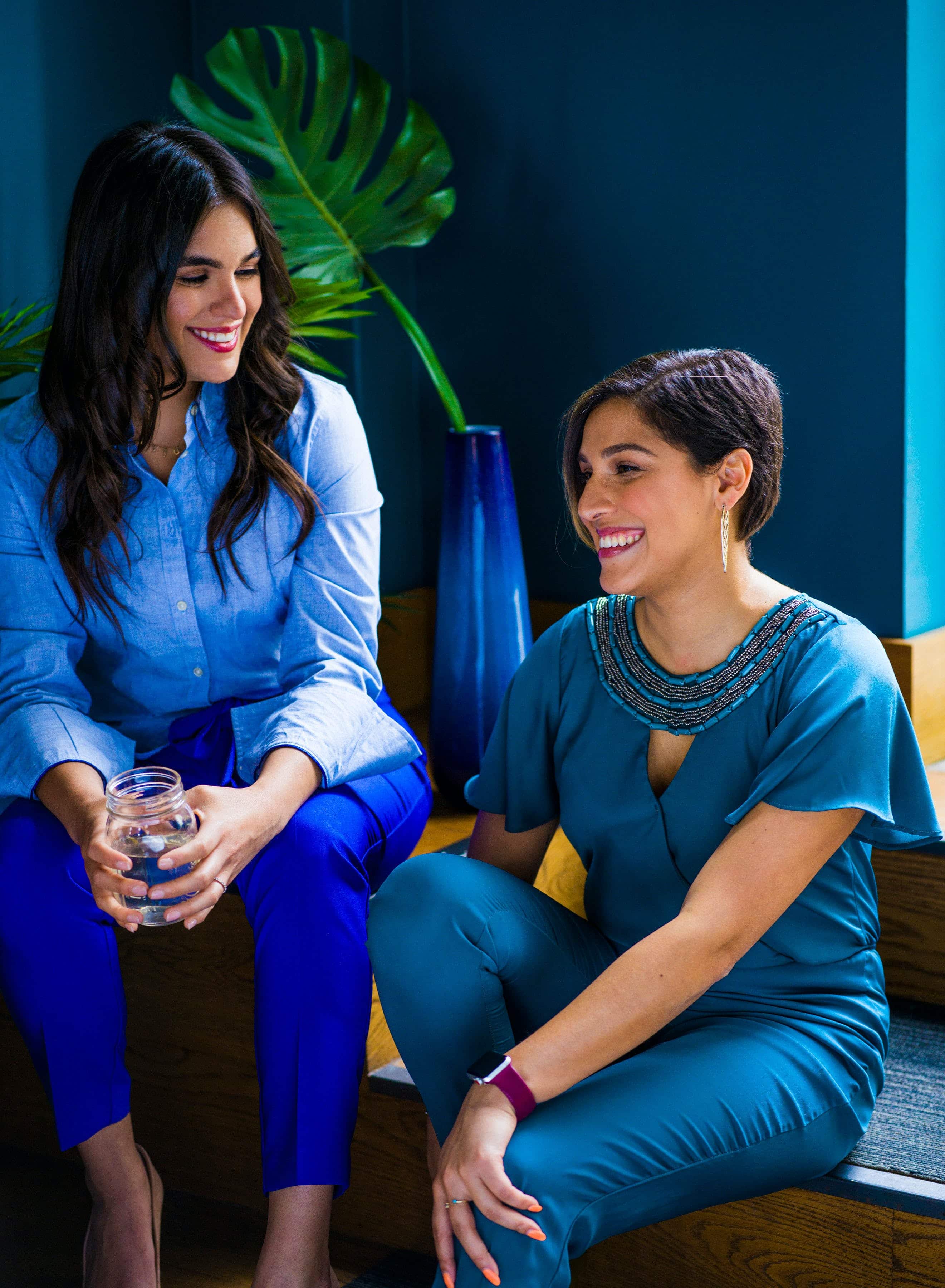 Two South Asian women, both wearing various shades of blue, sitting on furniture in front of a plant and a teal wall, talking.