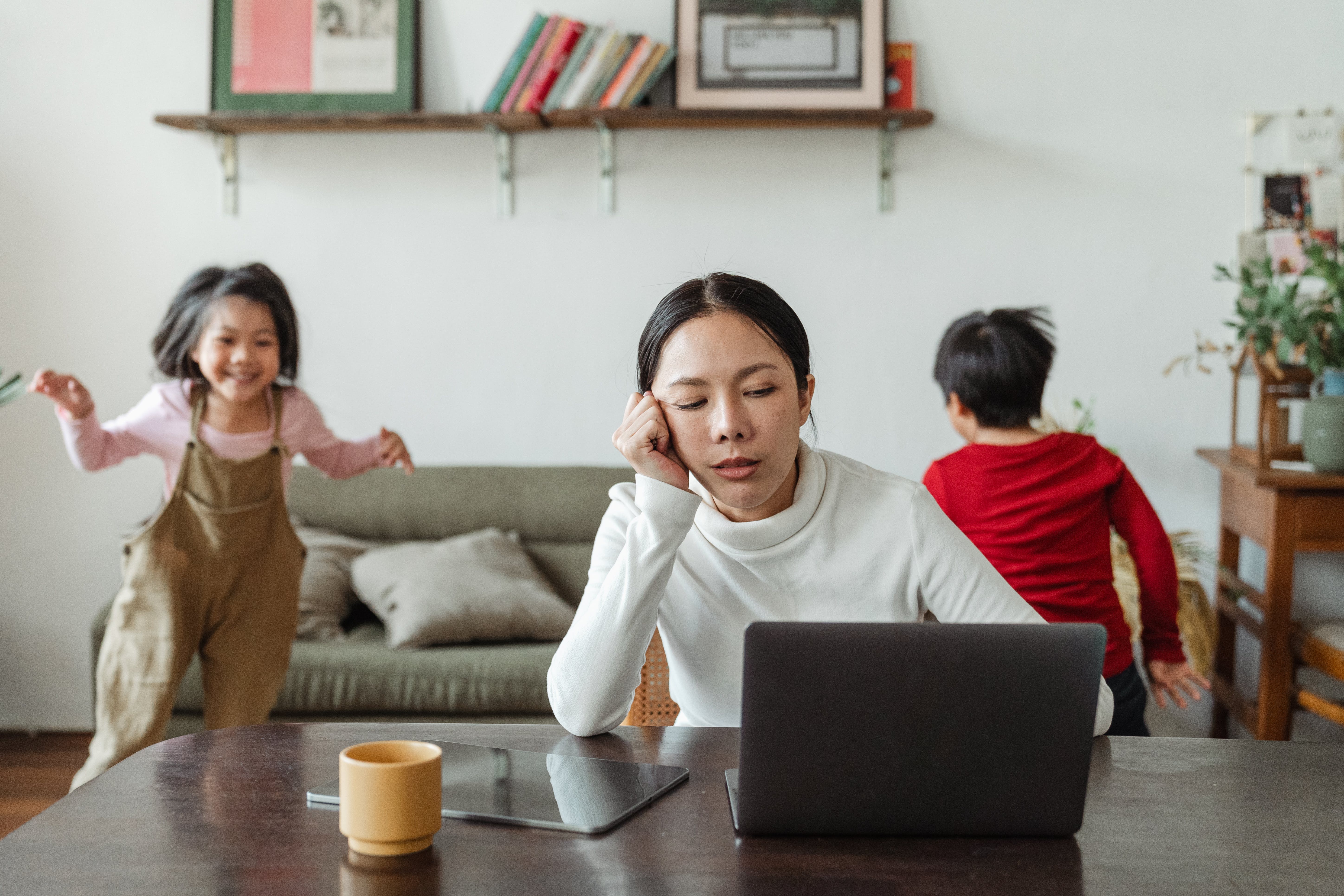 A woman sitting at her laptop looking stressed, while kids run in the background