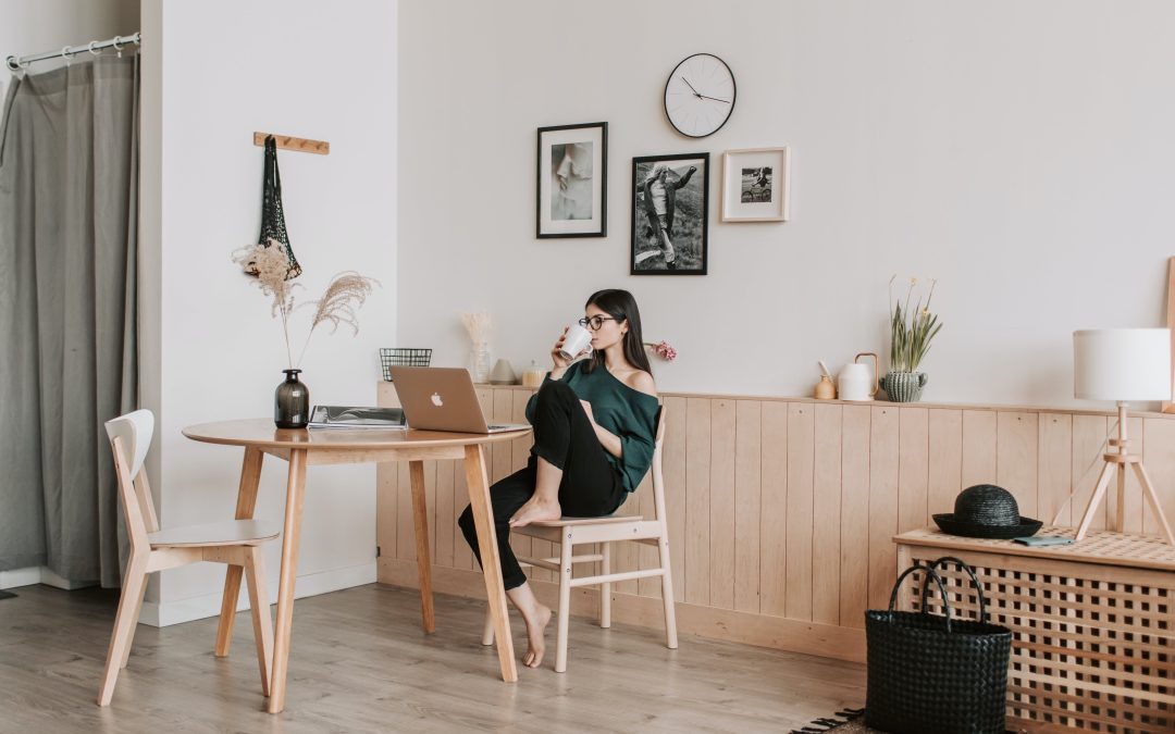 A white woman sitting at a table sipping from a mug, looking at a laptop