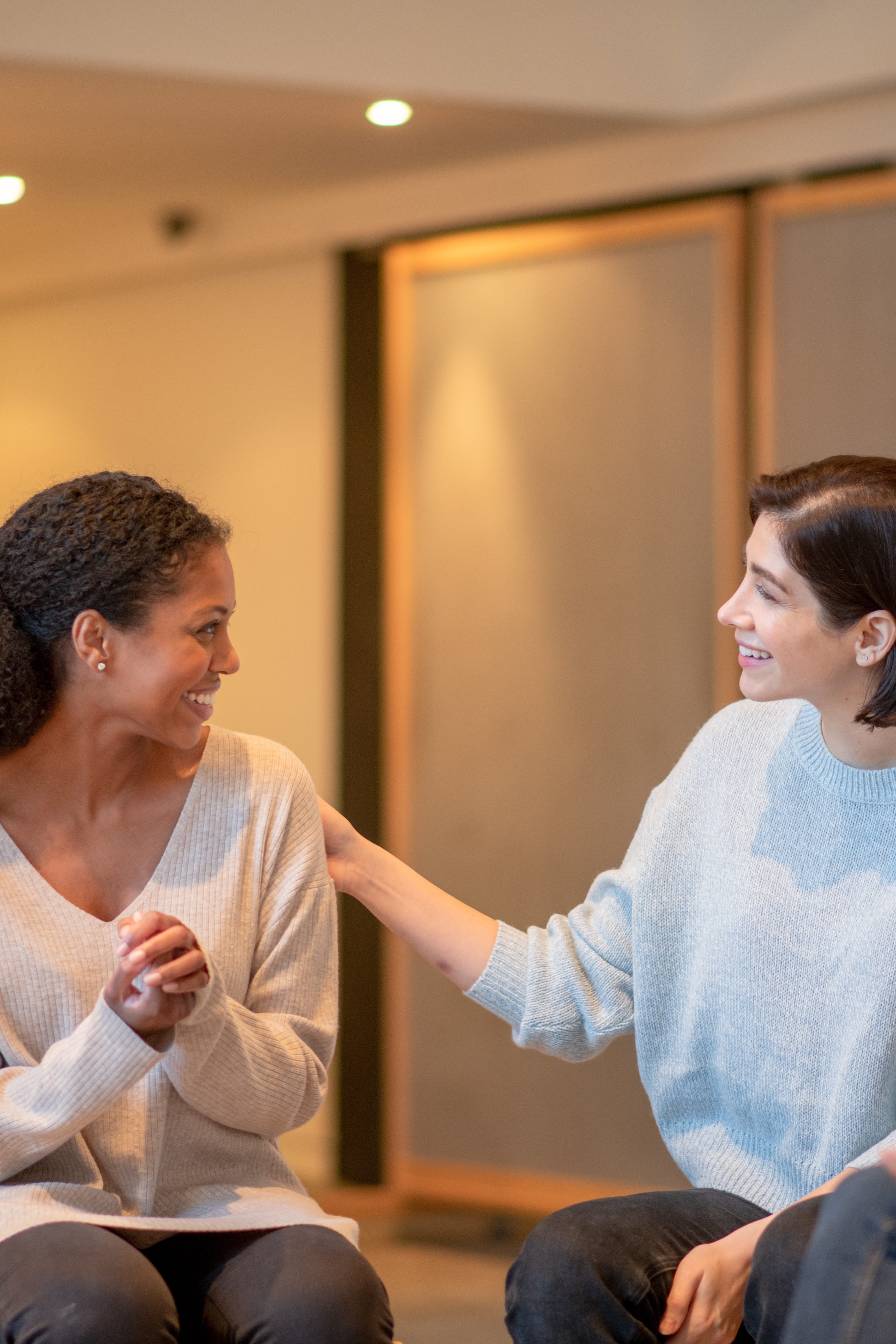 A young Black woman sitting next to a young white woman, clasping her hands with the white woman's hand on her shoulder in a show of support. They are smiling at each other.