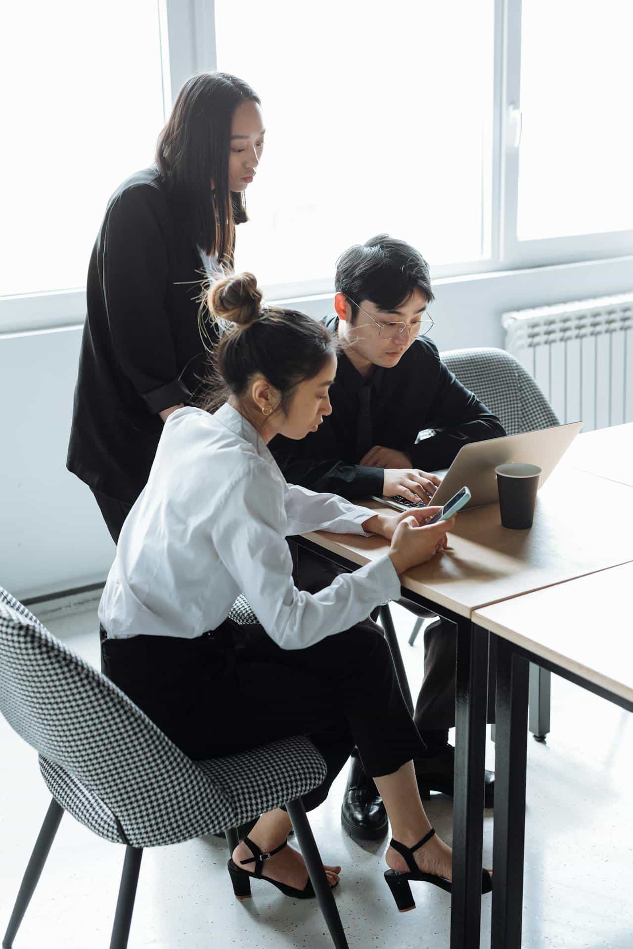 A group of three Asian American corporate professional workers sitting at a table and looking at their phones/laptops.