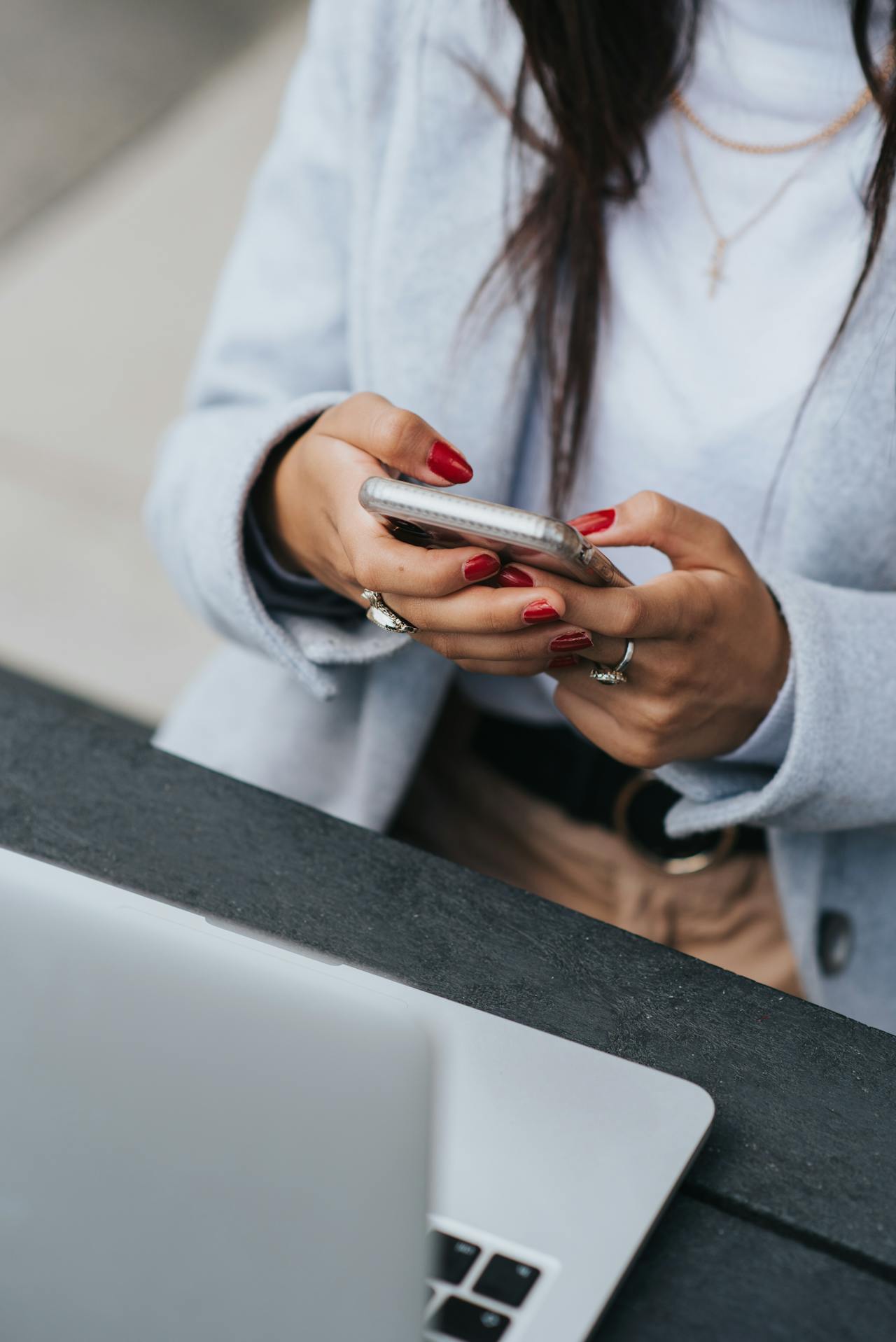 Close cropped photo of a woman in a business casual suit and red manicure working on her phone, with part of her laptop visible in the foreground. Only her chest and arms are visible in the photo.