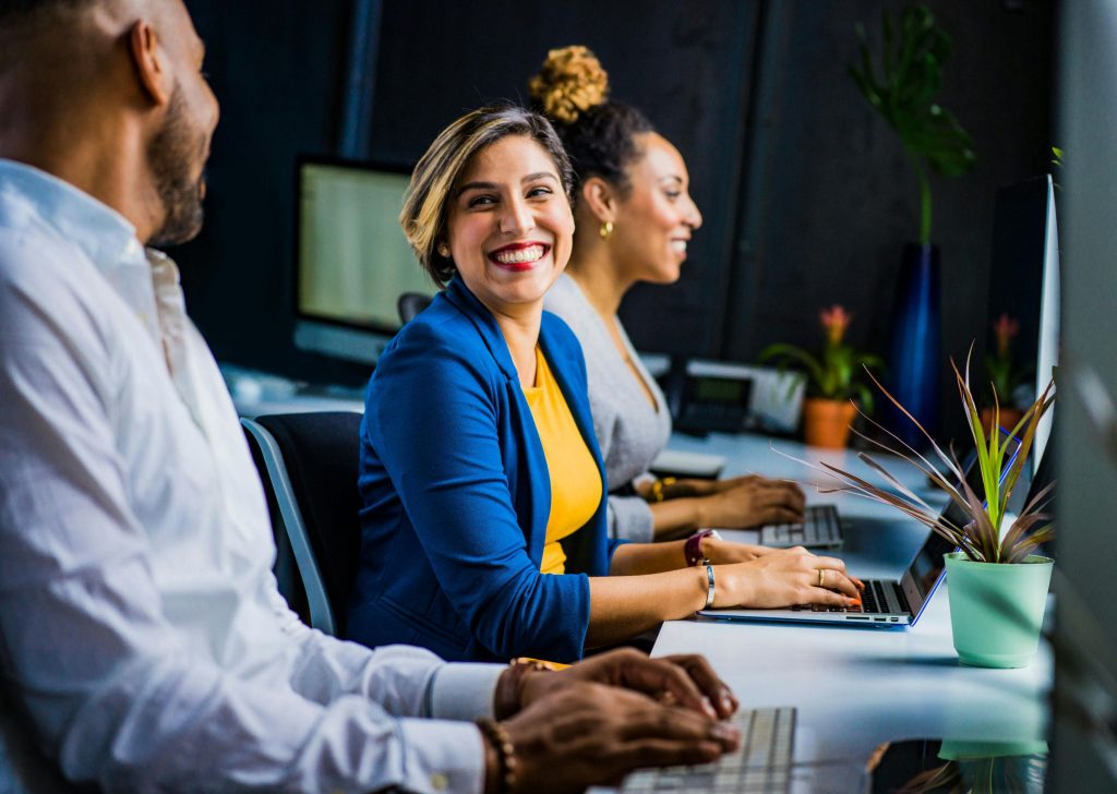 A woman with brown skin and short dark hair with blonde highlights, sitting at a laptop at a long desk between a Black woman, to her left, and a Black man, to her right. She is wearing red lipstick and smiling as she types.