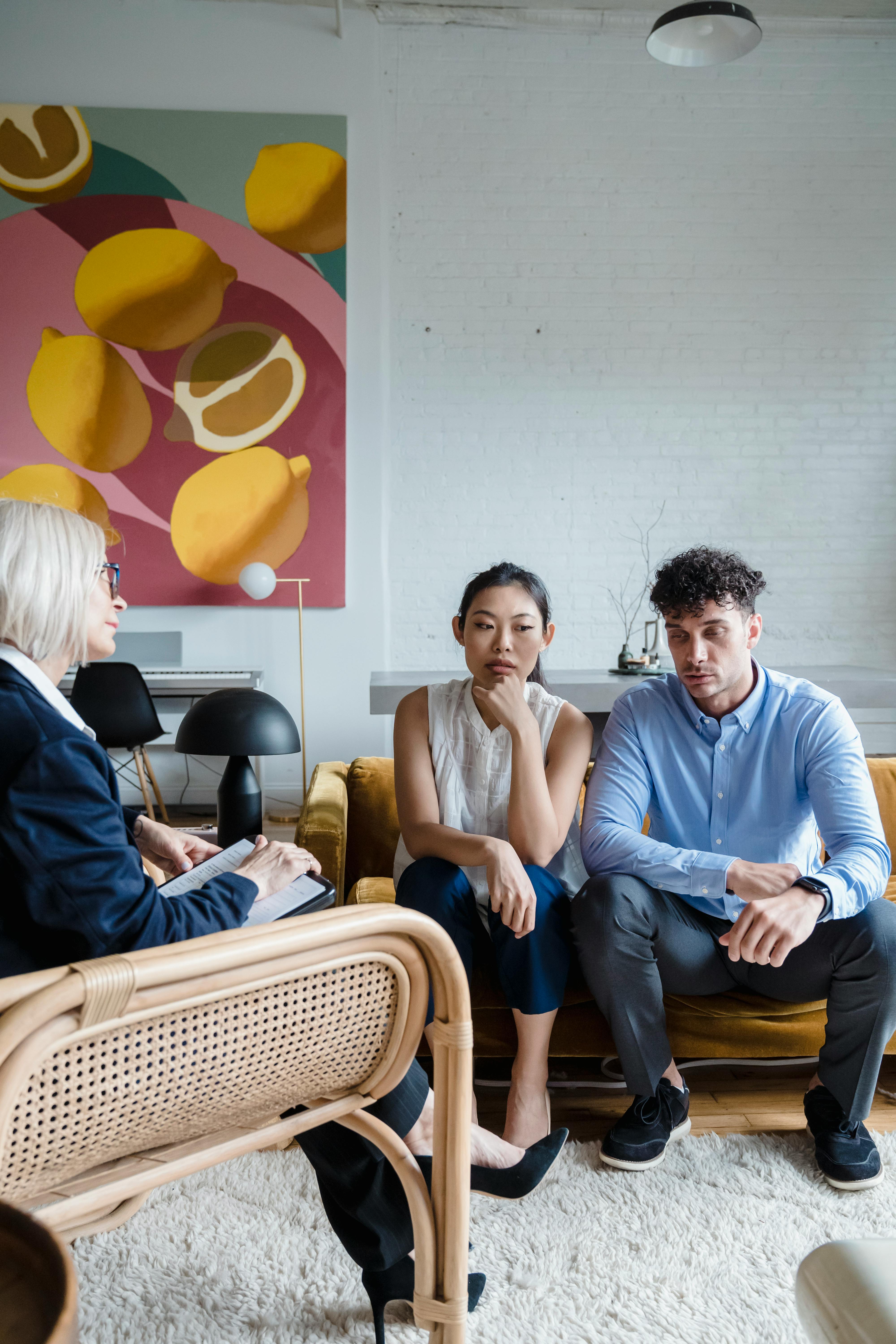 A white man and an Asian woman sitting next to one another on a couch with a white woman therapist slightly out of focus. 