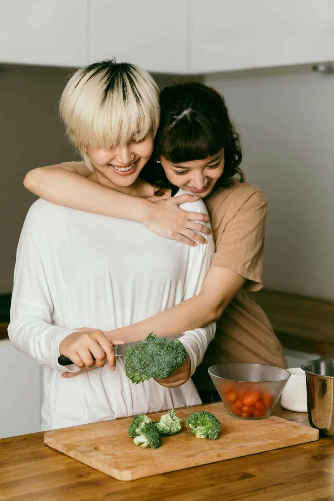 Two femme presenting, AAPI people are preparing a meal together and are being physically affectionate towards eath other. 