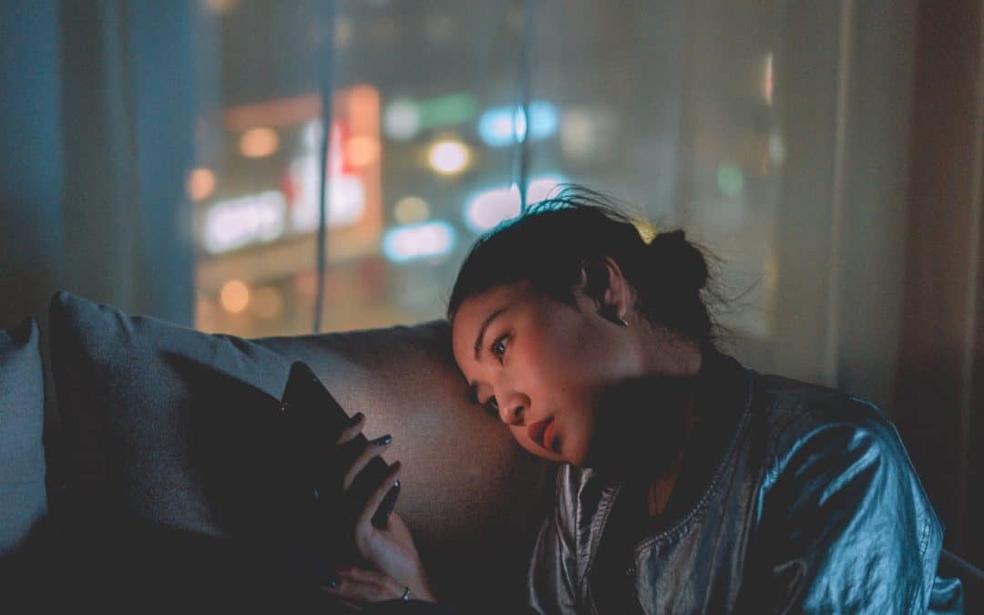 Woman sitting on a couch in the dark, looking absentmindedly at her phone.