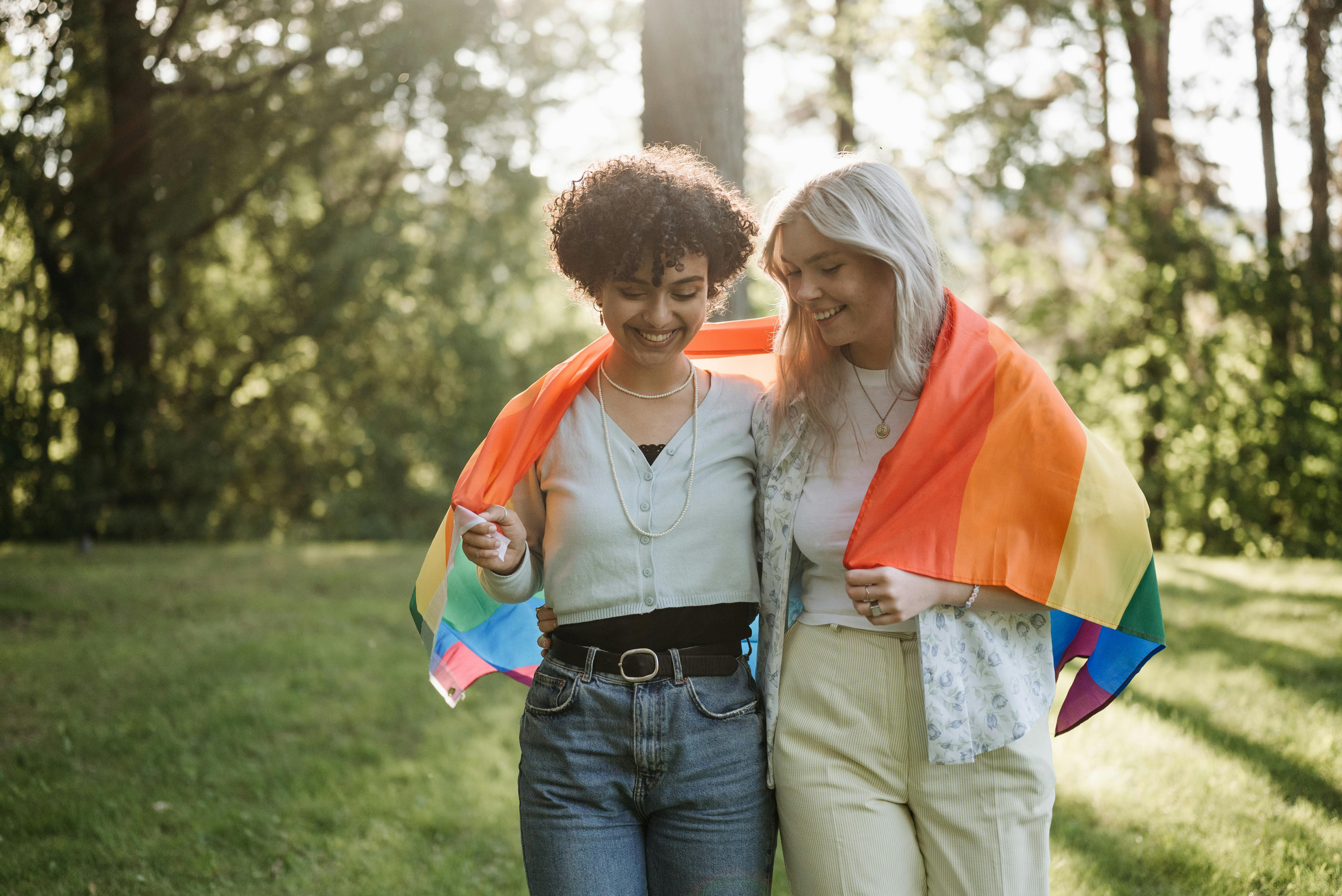 A queer woman of color and a white queer woman laughing and walking together with a Pride flag draped over their shoulders. 