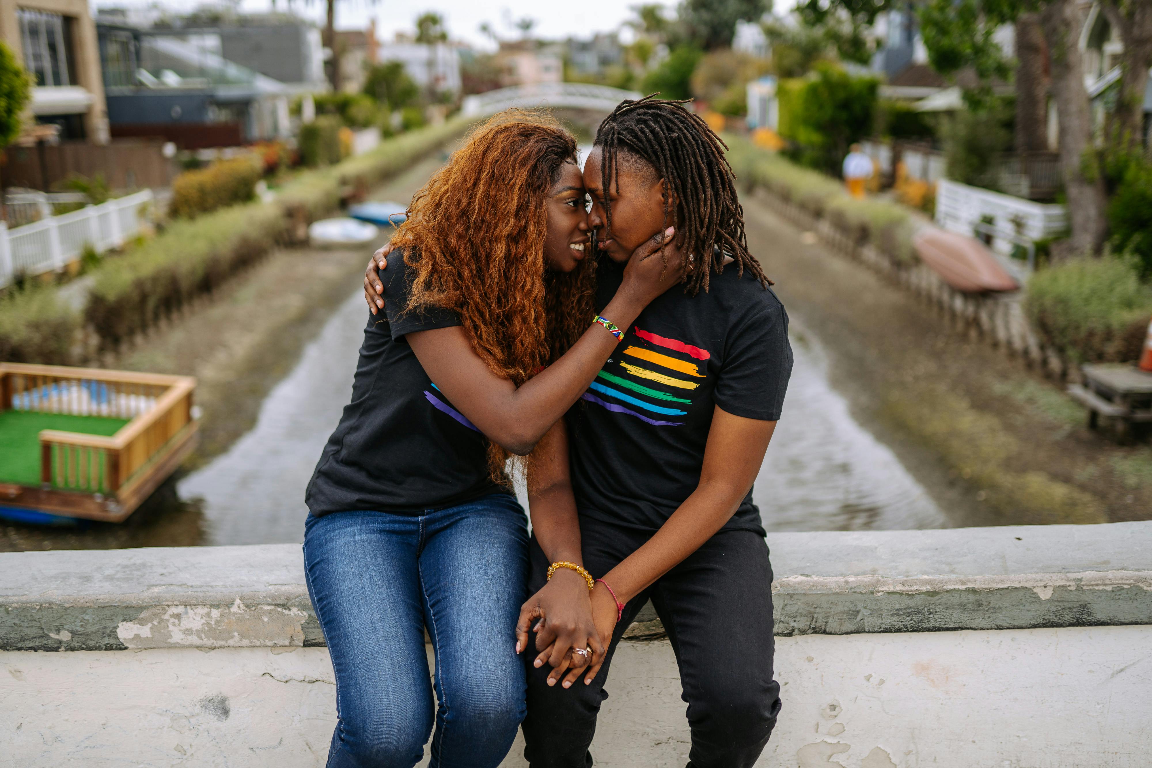 Two Black queer women sitting together in Pride shirts; one holding the other's face in joy. 