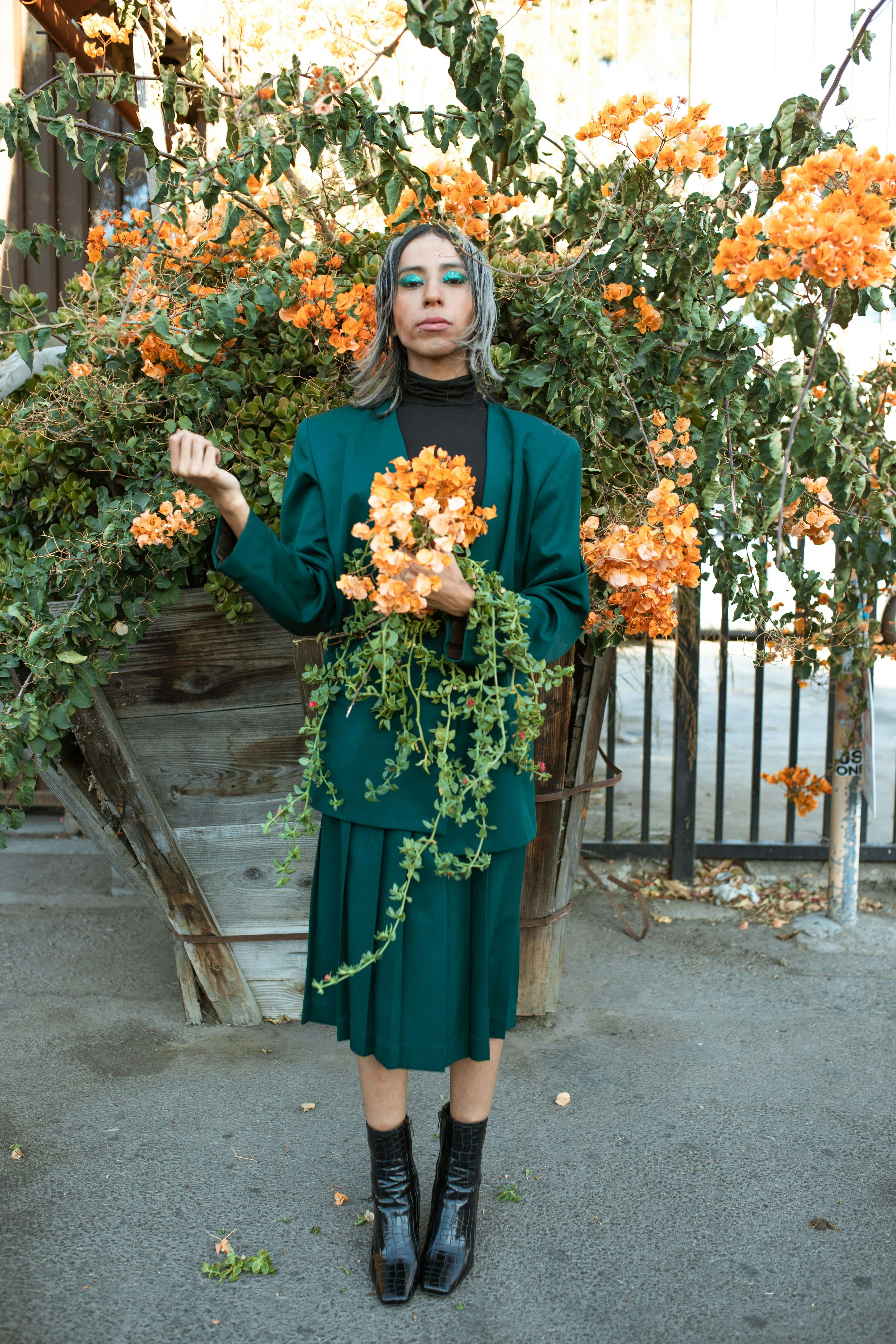 A femme presenting person with sparkly eyeshadow and a green suit poses outdoors with flowers.