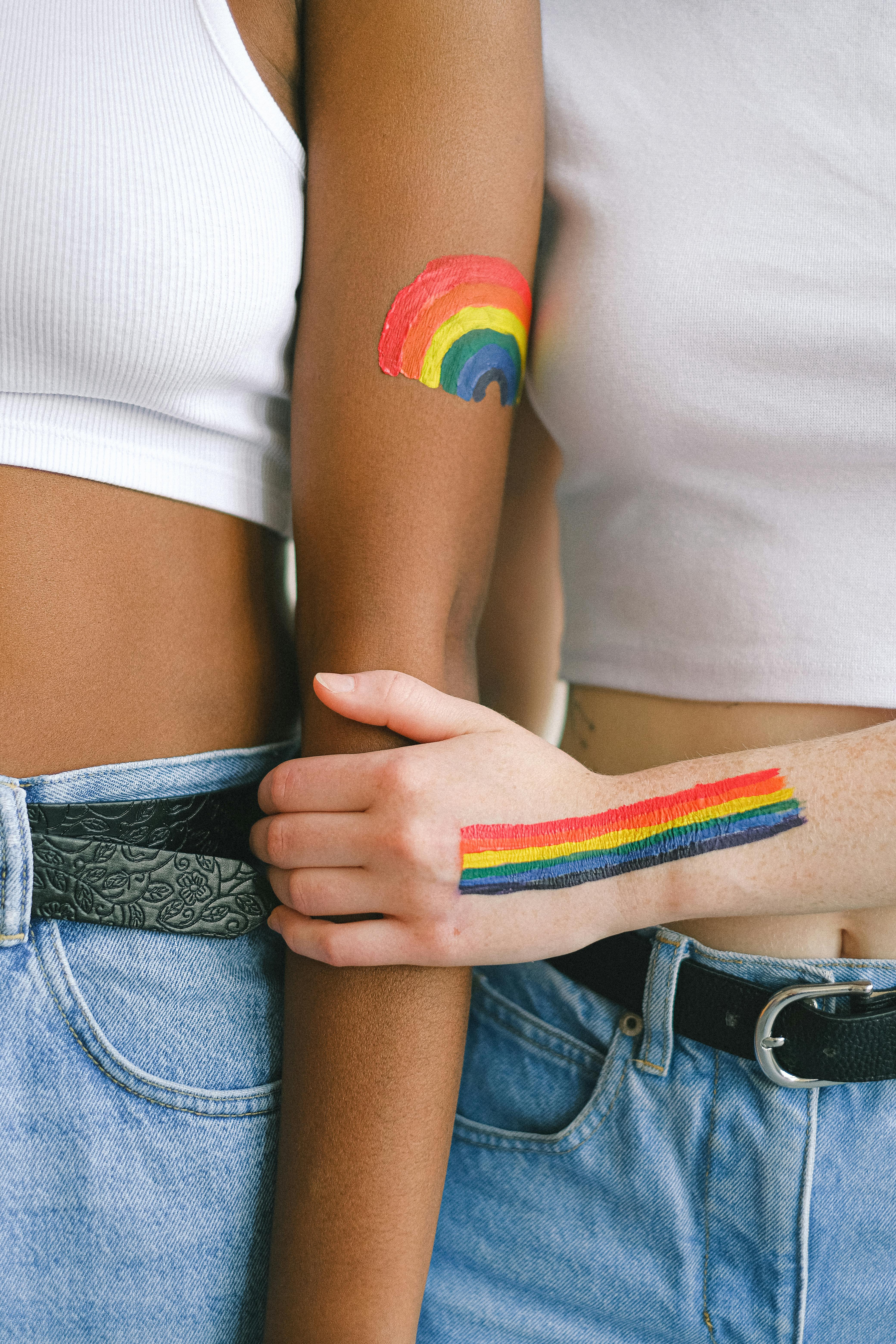 Queer women's arms next to one another, both painted with rainbow flags or rainbows; the white women's hand is holding the Black woman's arm in affection. 