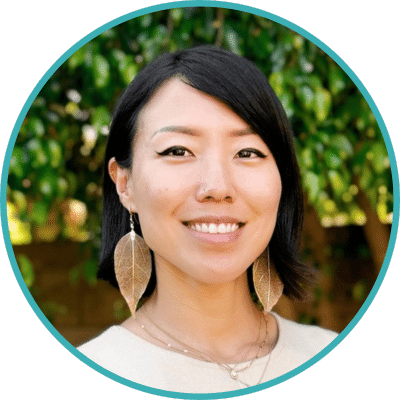 Outdoor headshot of Esther Kwon, an East Asian woman with a bob haircut, wearing gold dipped leaf earrings and smiling towards the camera.