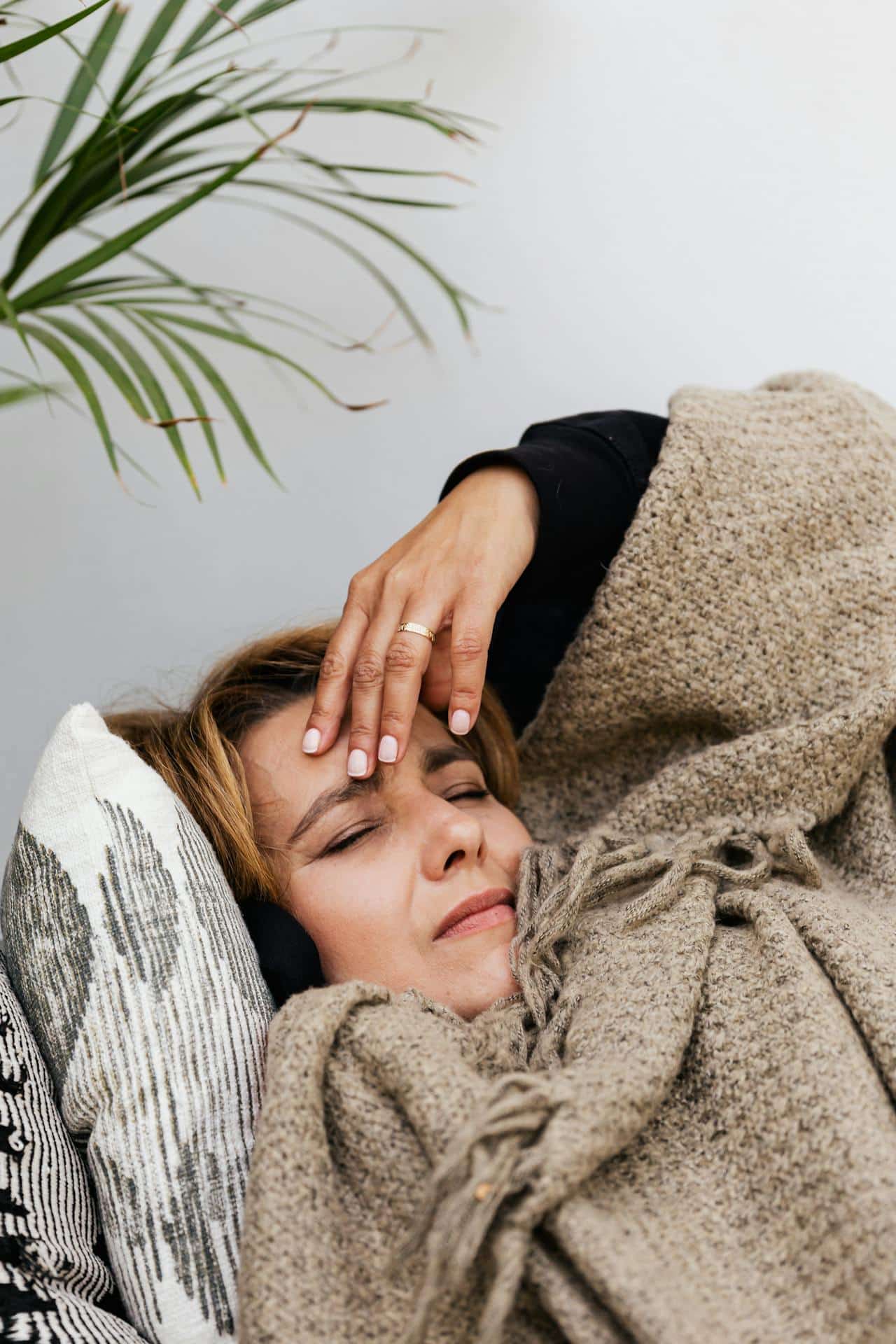 A woman rests on the sofa under a blanket, with her hand above her head as though she has a headache.