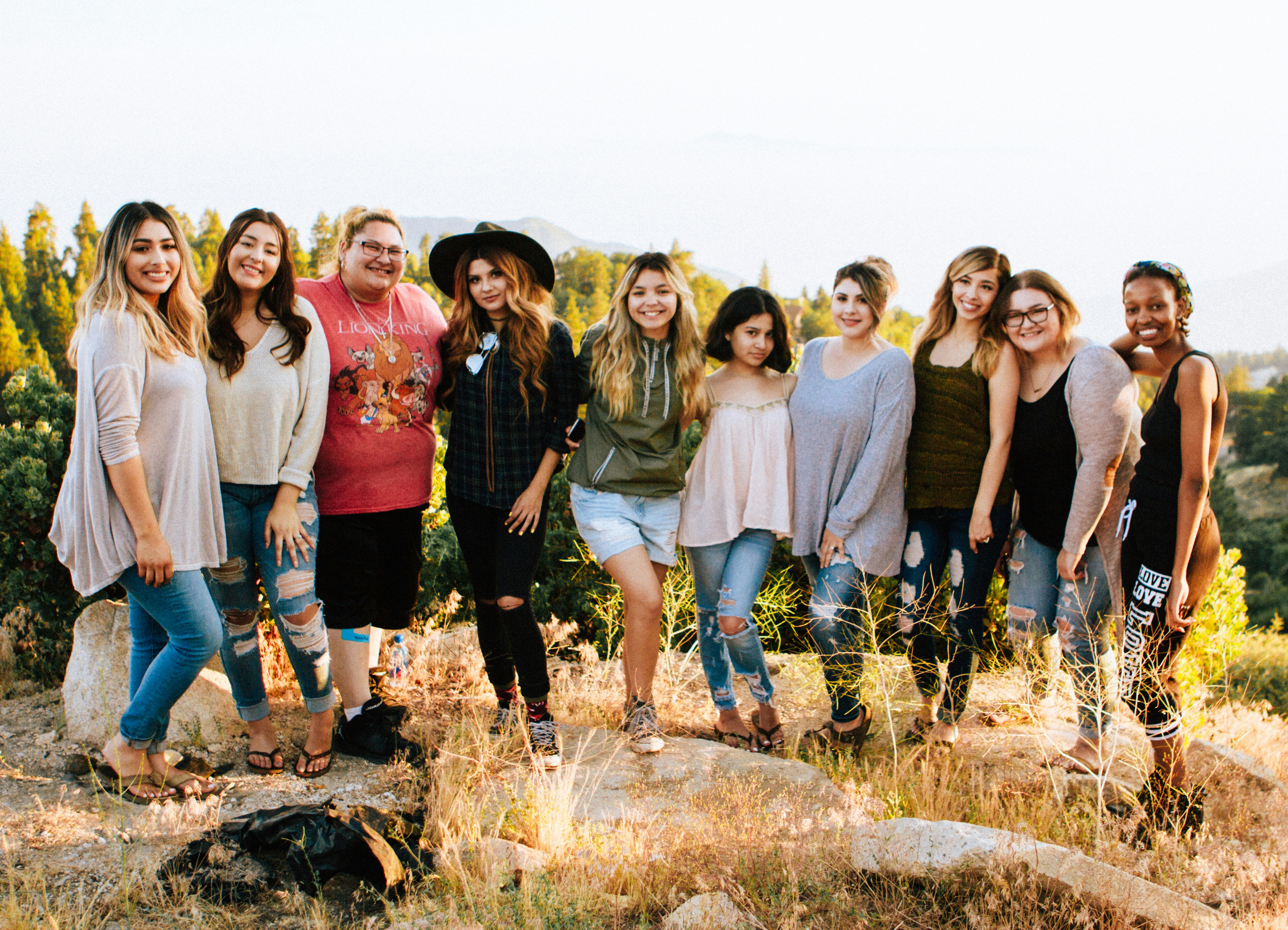 A group of 10 individuals of varying genders, races, and sizes, standing together smiling outside on a rocky overlook, with mountains distantly in the background. 