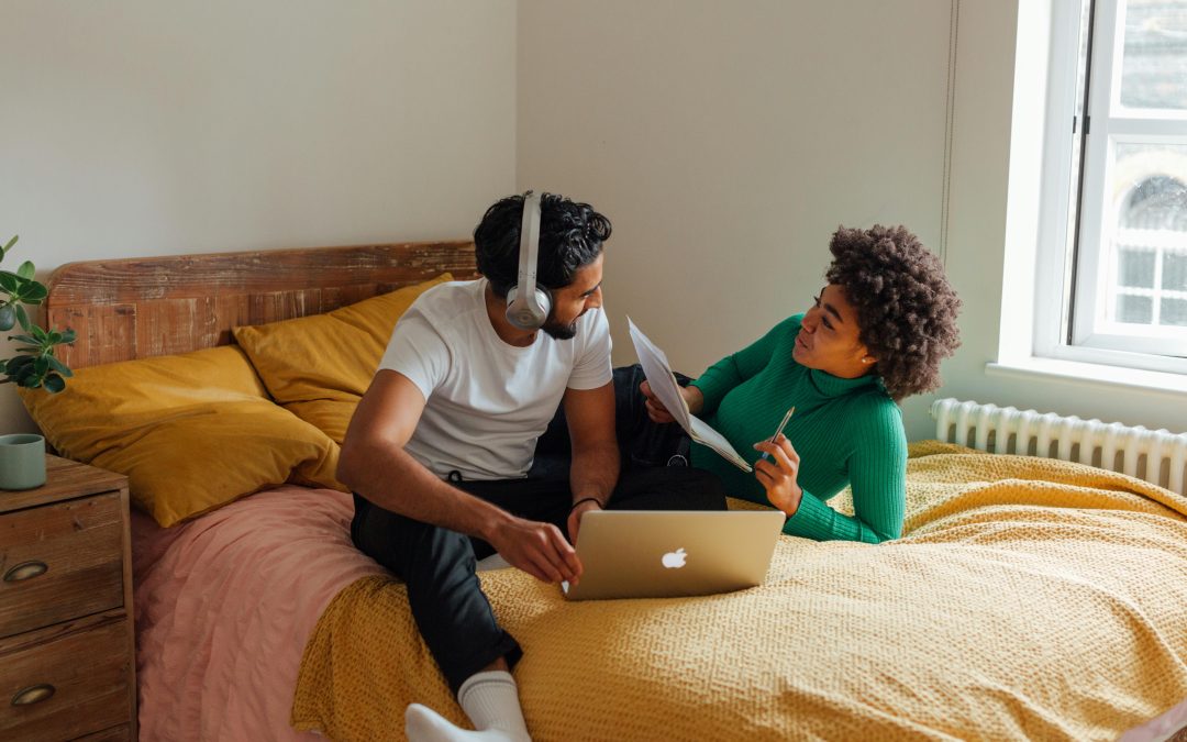 A Black woman and a man of South Asian heritage sitting together on a bed with yellow linens, talking to each other. The woman, on the right, is holding a pile of papers and a pen, and the man is wearing over ear headphones and has a laptop in front of him.