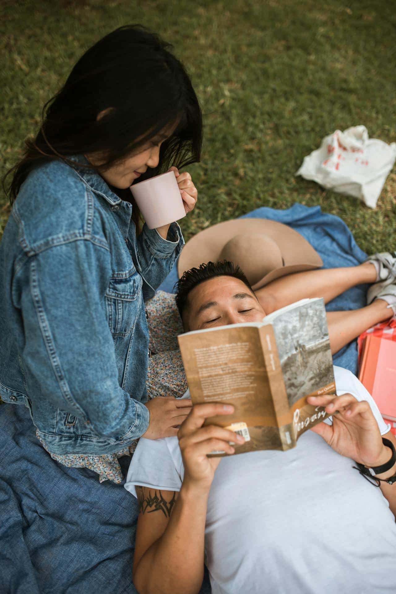 A bisexual couple, an Asian man and woman, relaxing in a park reading and drinking coffee.