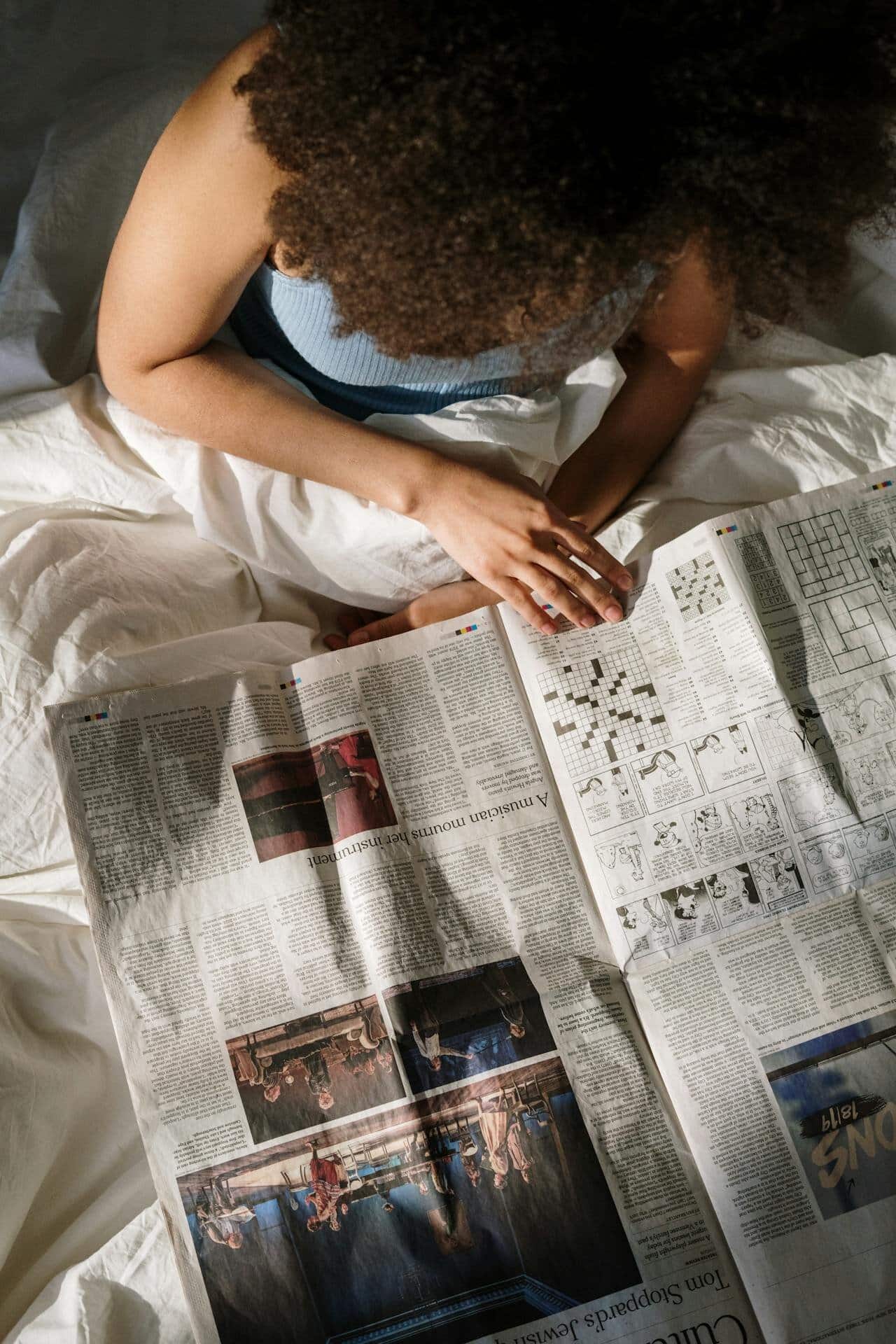 An overhead shot of a black woman with a dark afro sitting under her sheets reading a newspaper.