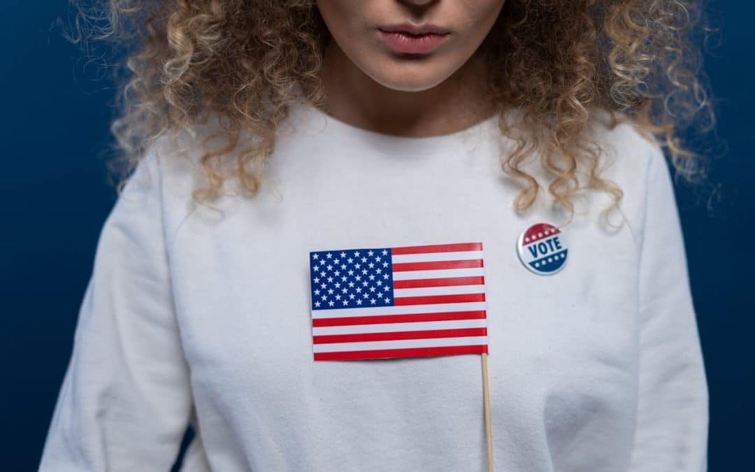 Half of a woman's face is showing, her skin is medium toned and she has blonde curly hair. She's wearing a white long-sleeve and holding a small USA flag in front of her.