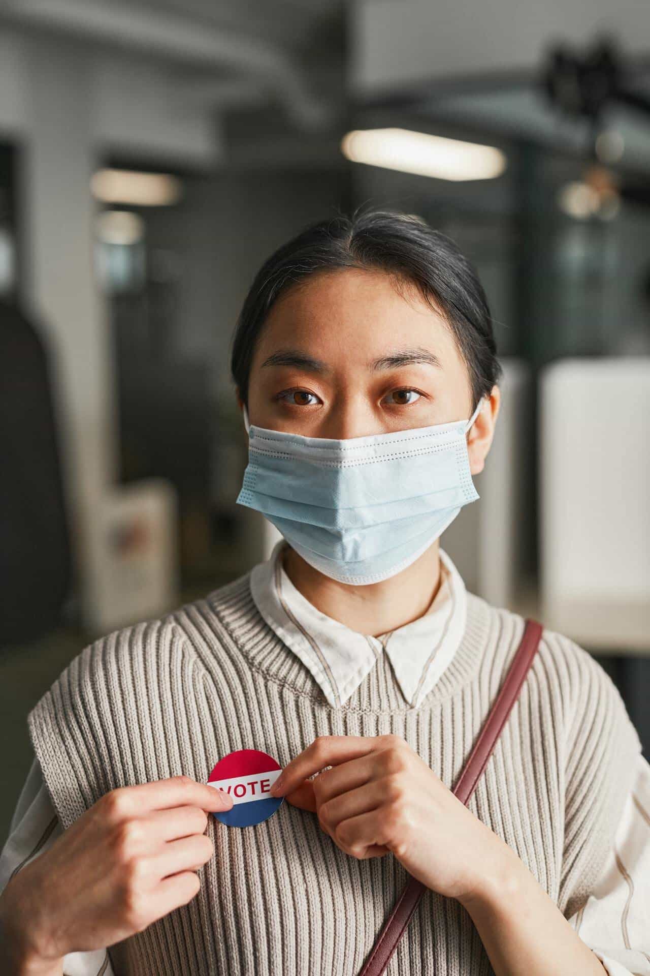 An Asian woman with a ponytail wears a mask while holding a "I voted" sign in front of her chest. She's wearing a white collared shirt underneath a cream colored sweater. 