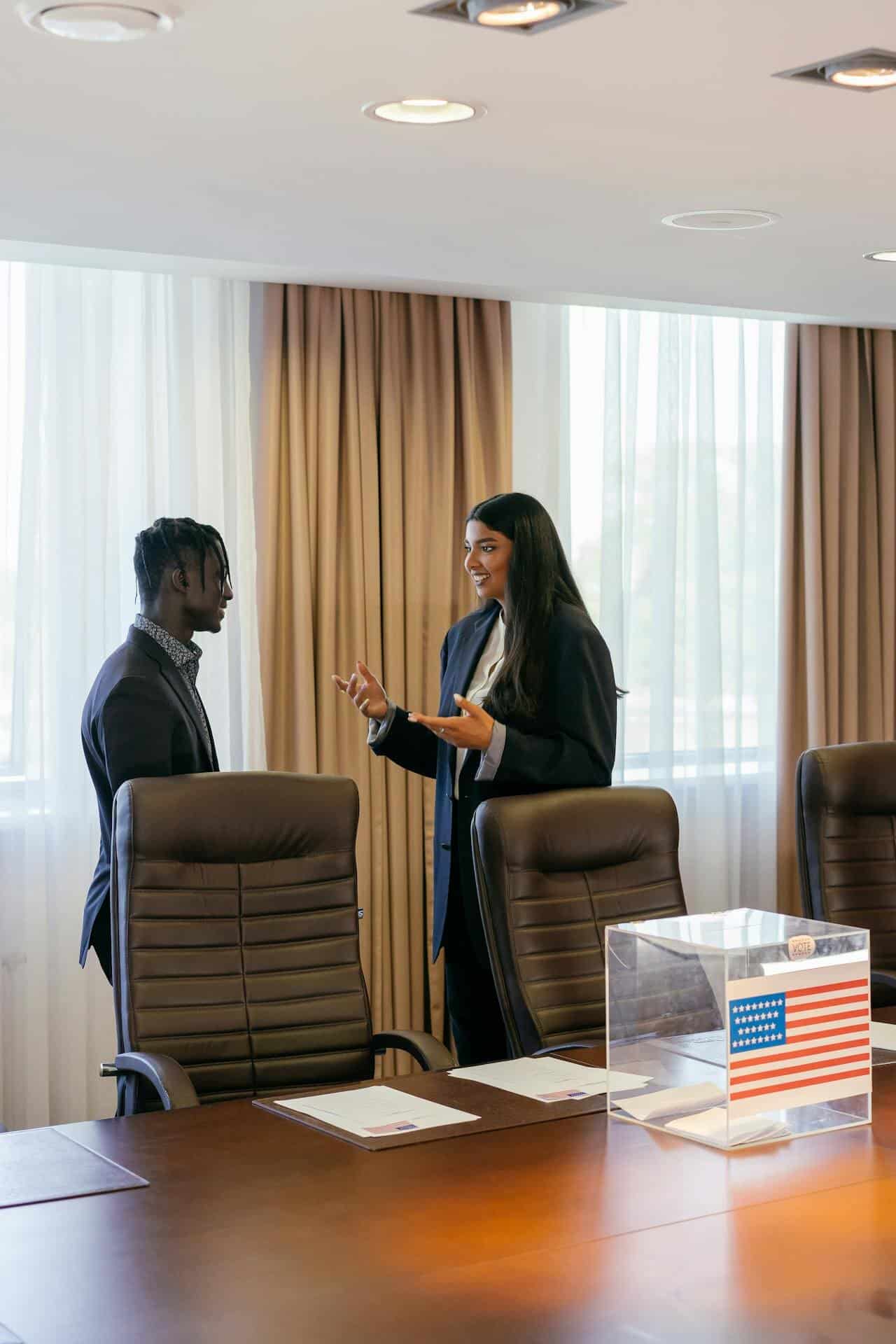 An Indian woman with long dark straight hair and dressed in a dark suit talks in a friendly manner to a black man dressed in a dark suit. They're standing in an office with a table, chairs, and a clear plastic box with an American flag on it in front of them. 